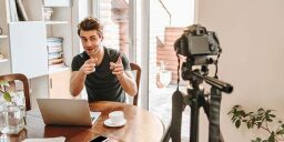 Person sitting at desk with laptop making finger guns at recording camera on a tripod