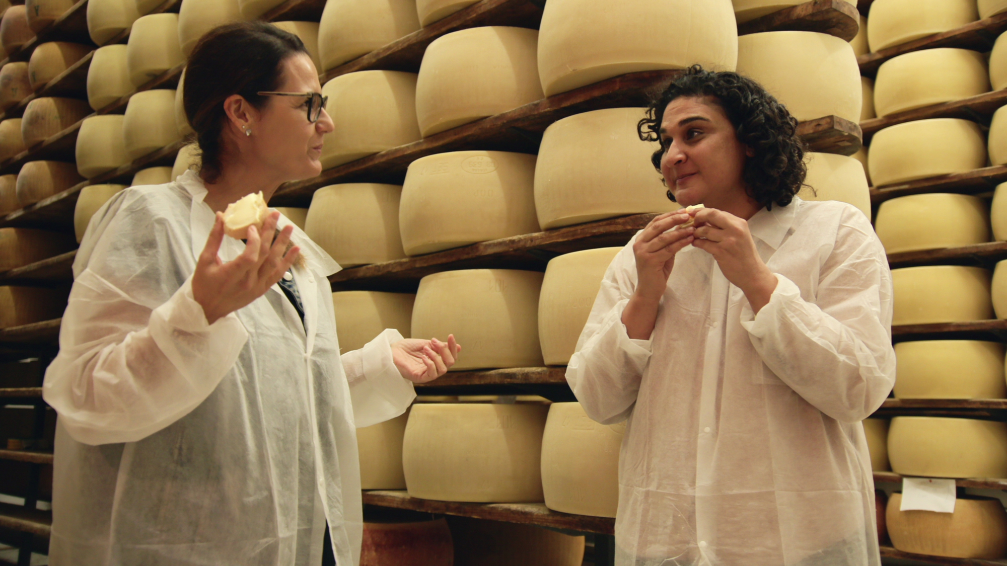 Two women, including host Samin Nosrat, eat cheese in front of a wall of cheese wheels