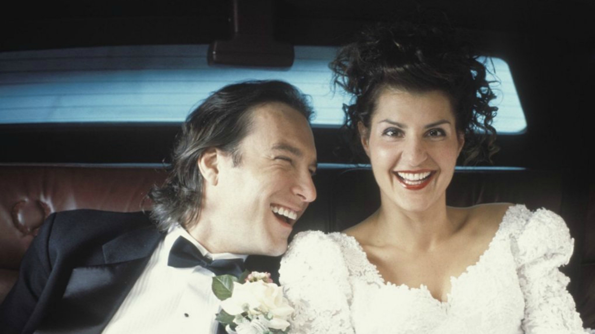 A woman in a wedding gown and a man in a tuxedo sit in the back seat of a car