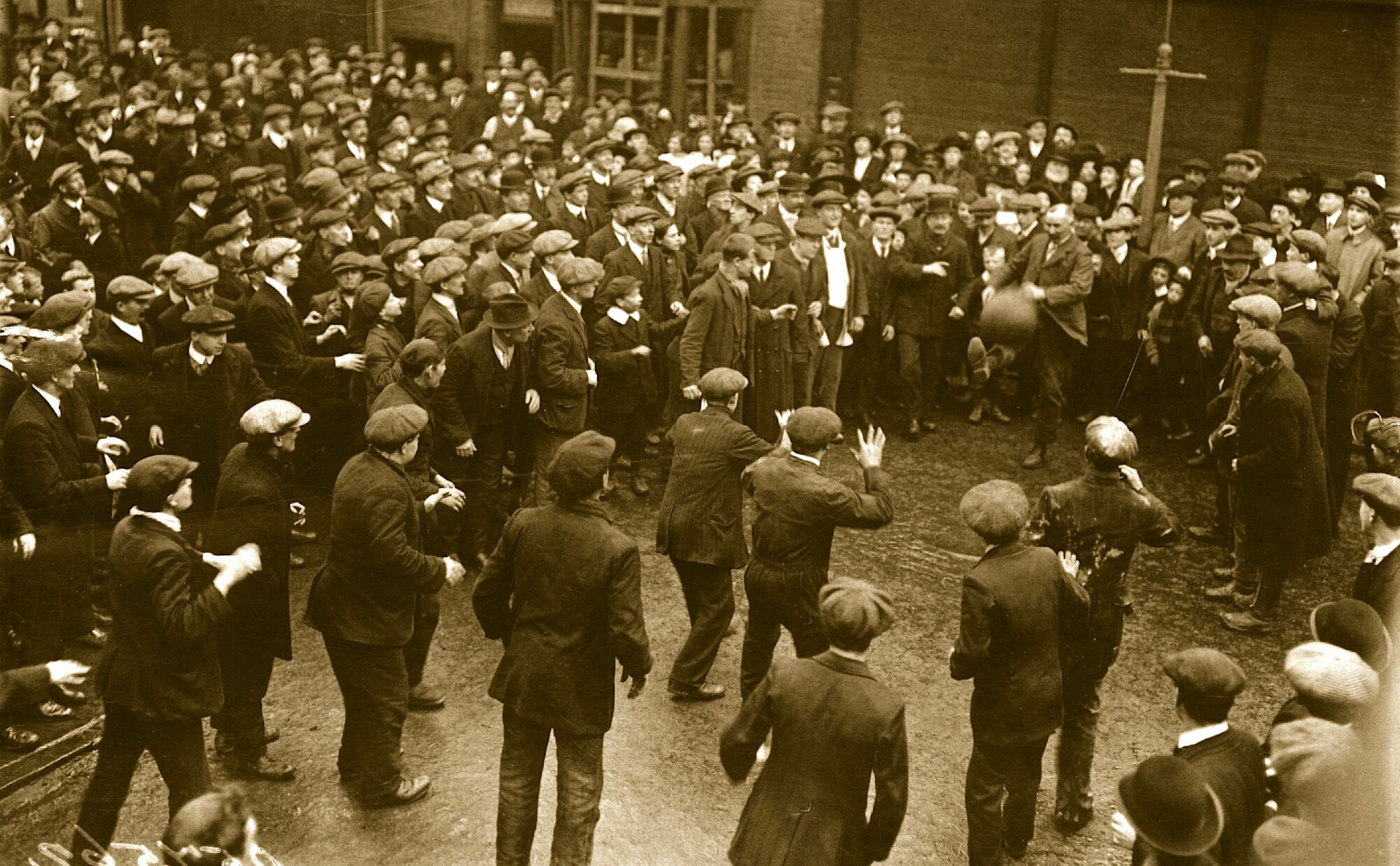 Men in flat caps stand around an Edwardian gentleman kicking a large ball to them.