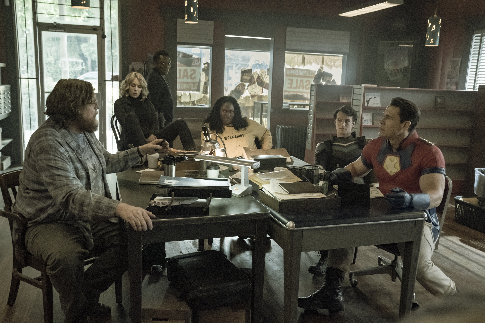 A group of individuals gathered around a table at a workspace; a still from "Peacemaker."