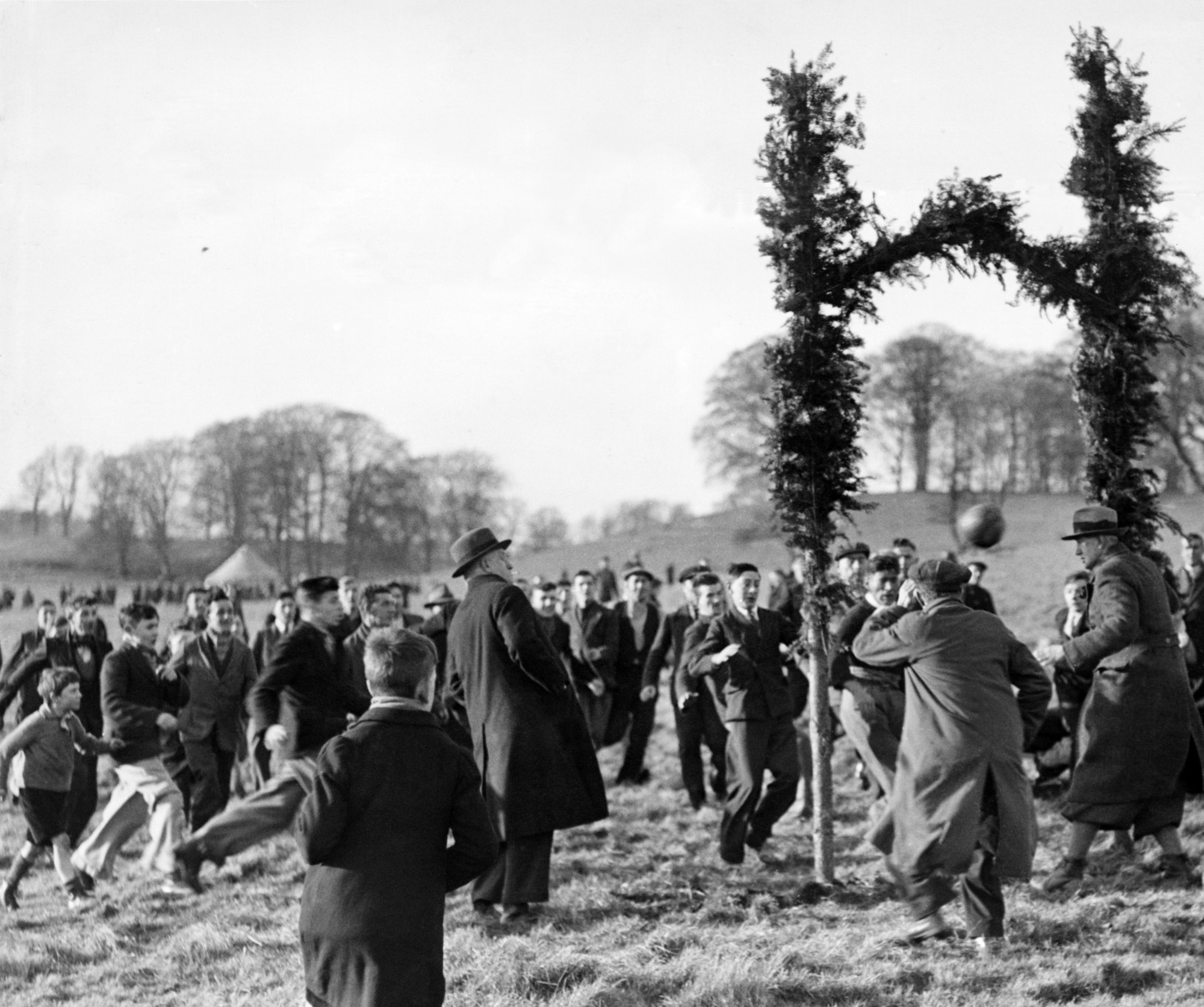 Men and boys kick a ball through a goal decorated with branches.