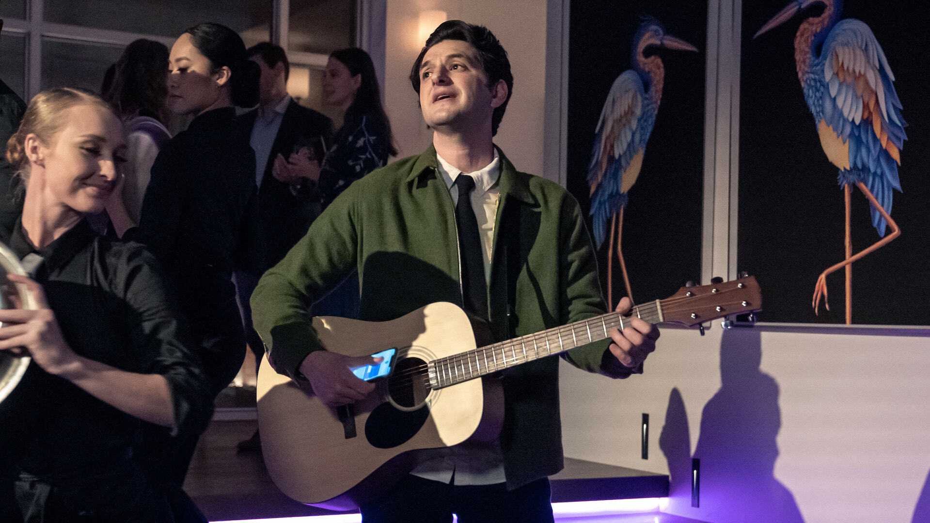 A man plays guitar in a fancy living room with bird portraits; a still from "The Afterparty."