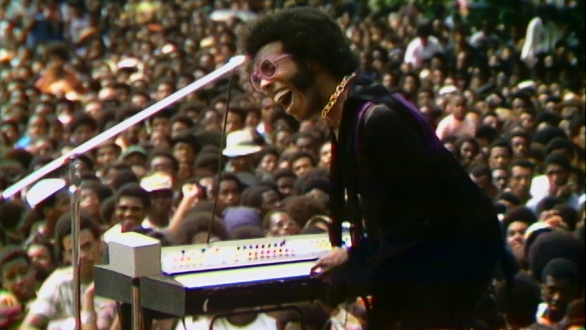 A black man sings into a microphone before a big concert crowd.
