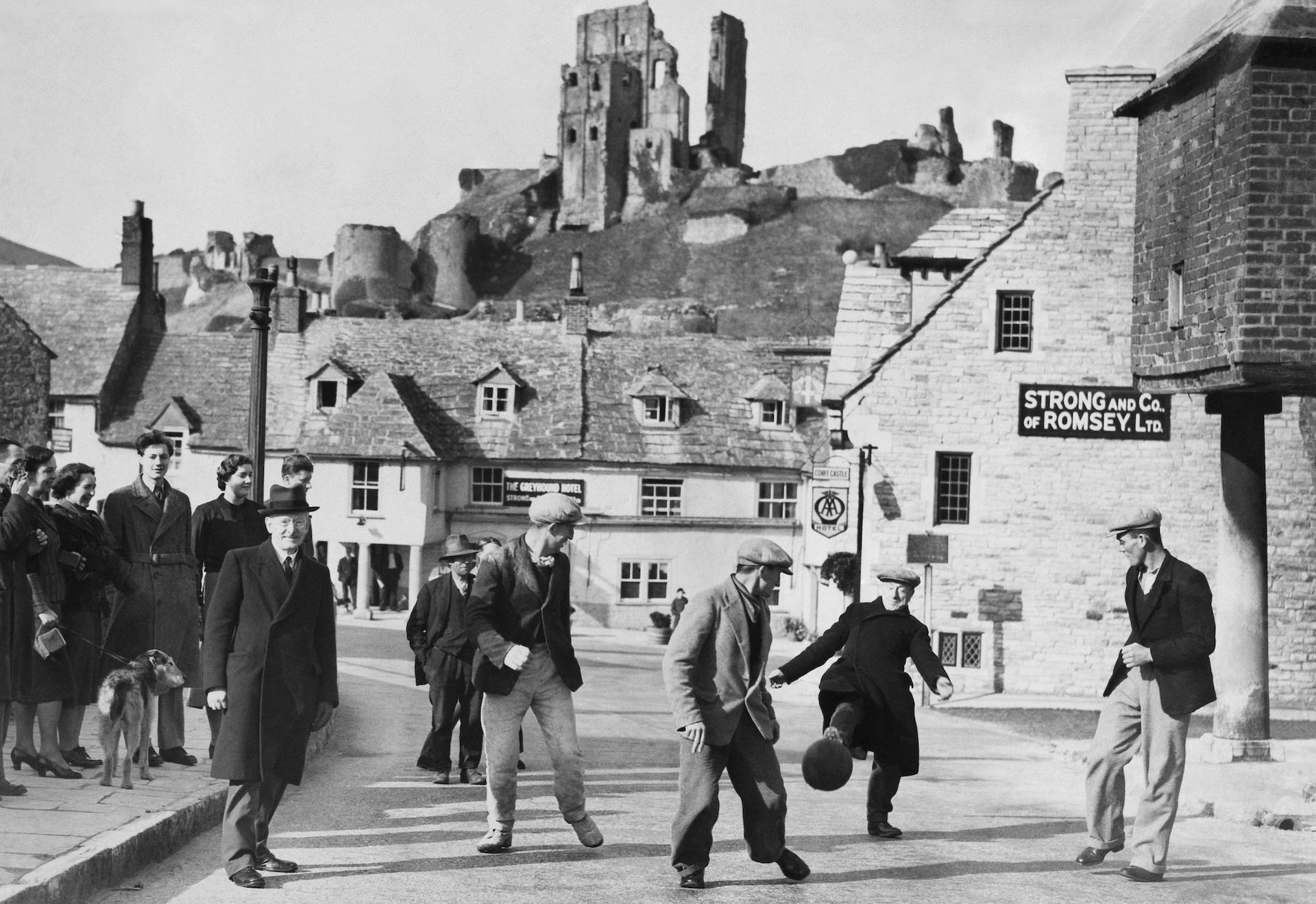 Men in flat caps kick a ball through the street of a town with a castle on a hill.