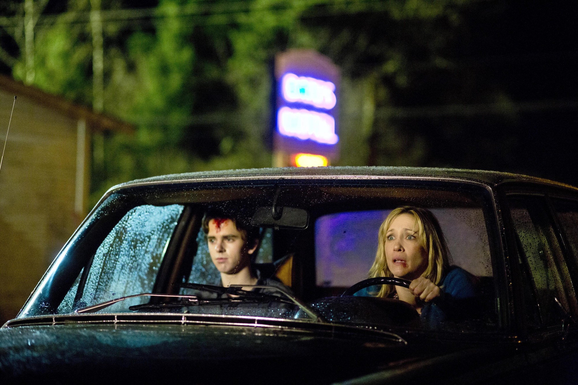A mother and son sit in e car in the rain. They look distressed and bloody. 