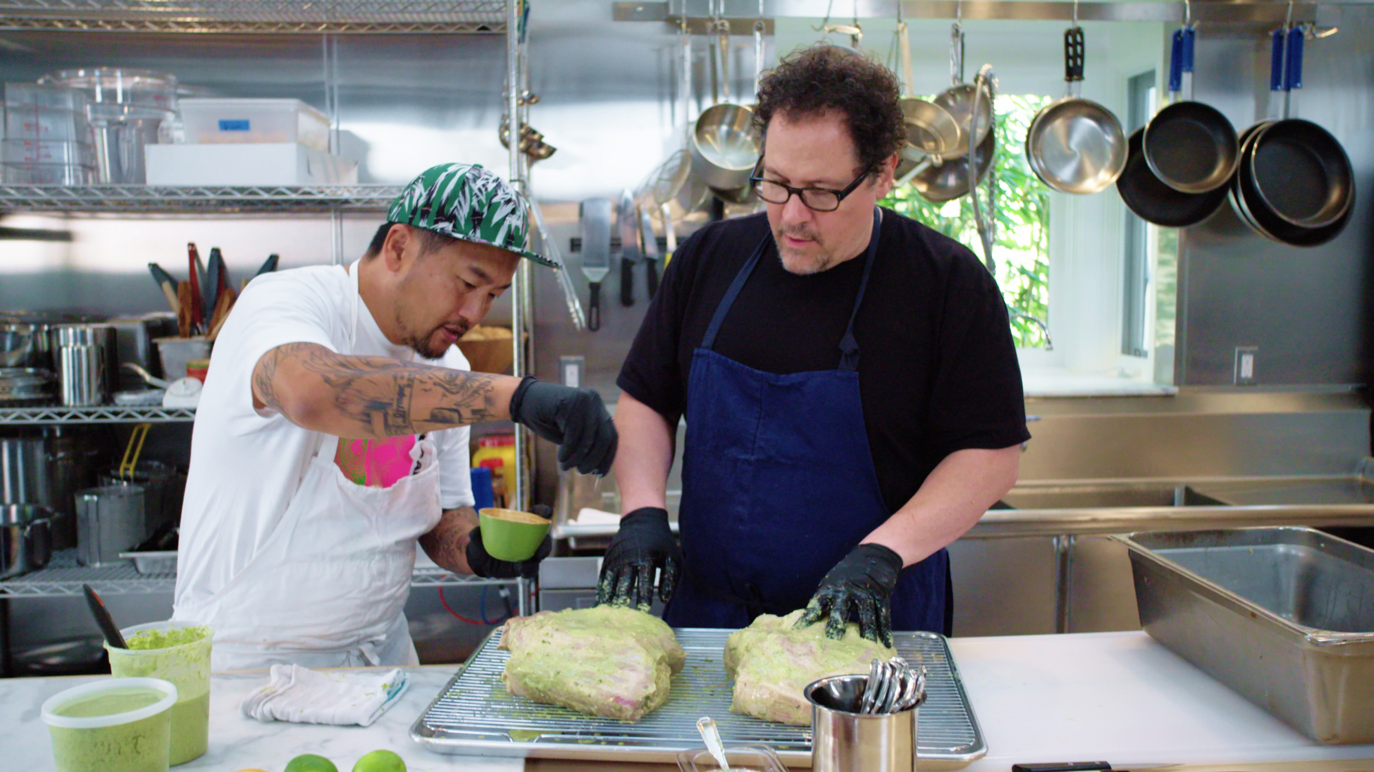 Two men, hosts Roy Choi and Jon Favreau, stand side by side behind a kitchen counter. They are seasoning two large pieces of meat.