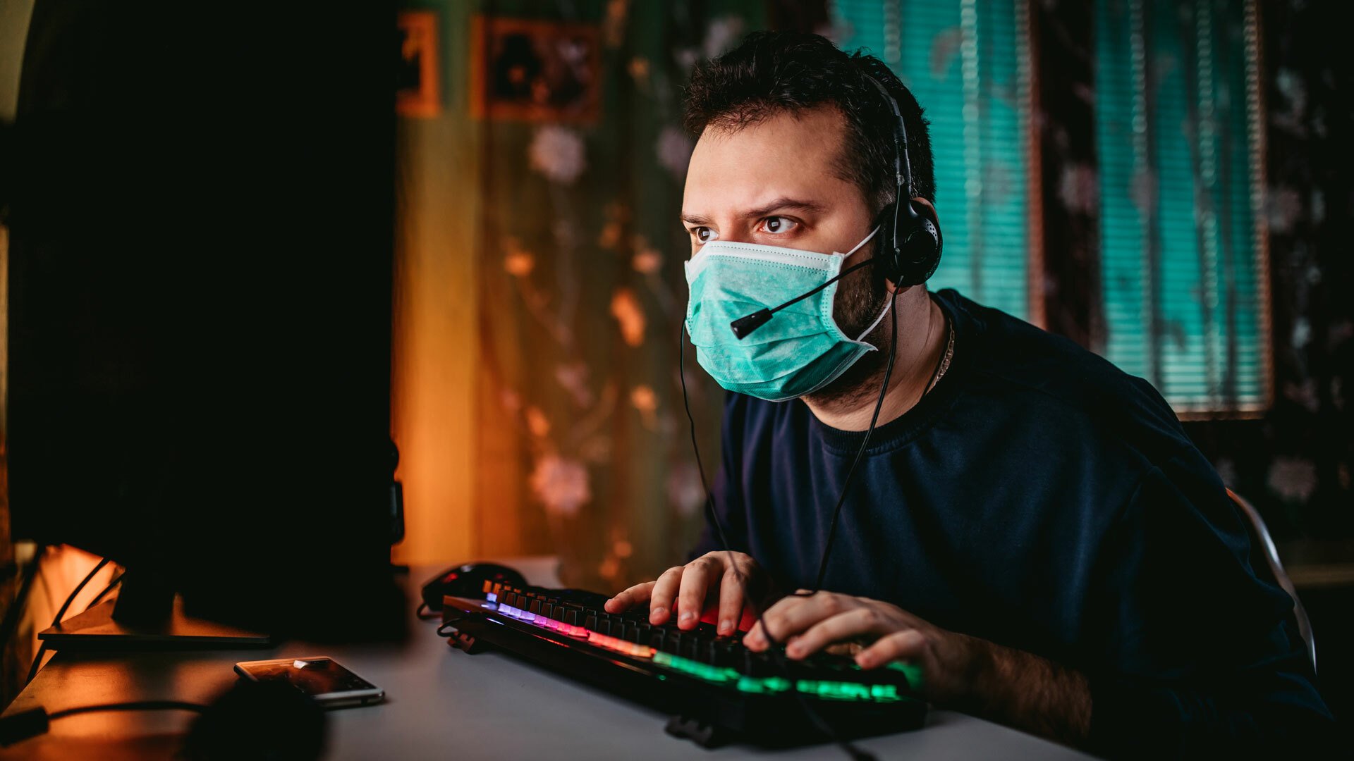 A man wearing a surgical mask and a mic-equipped gaming headset sits in front of a computer monitor and RGB-lit keyboard, leaning forward and staring at the screen intently.