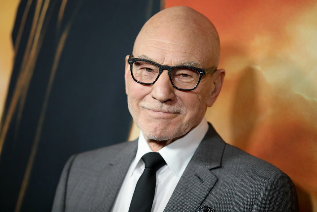 Sir Patrick Stewart, wearing thick black glasses and smiling slightly.