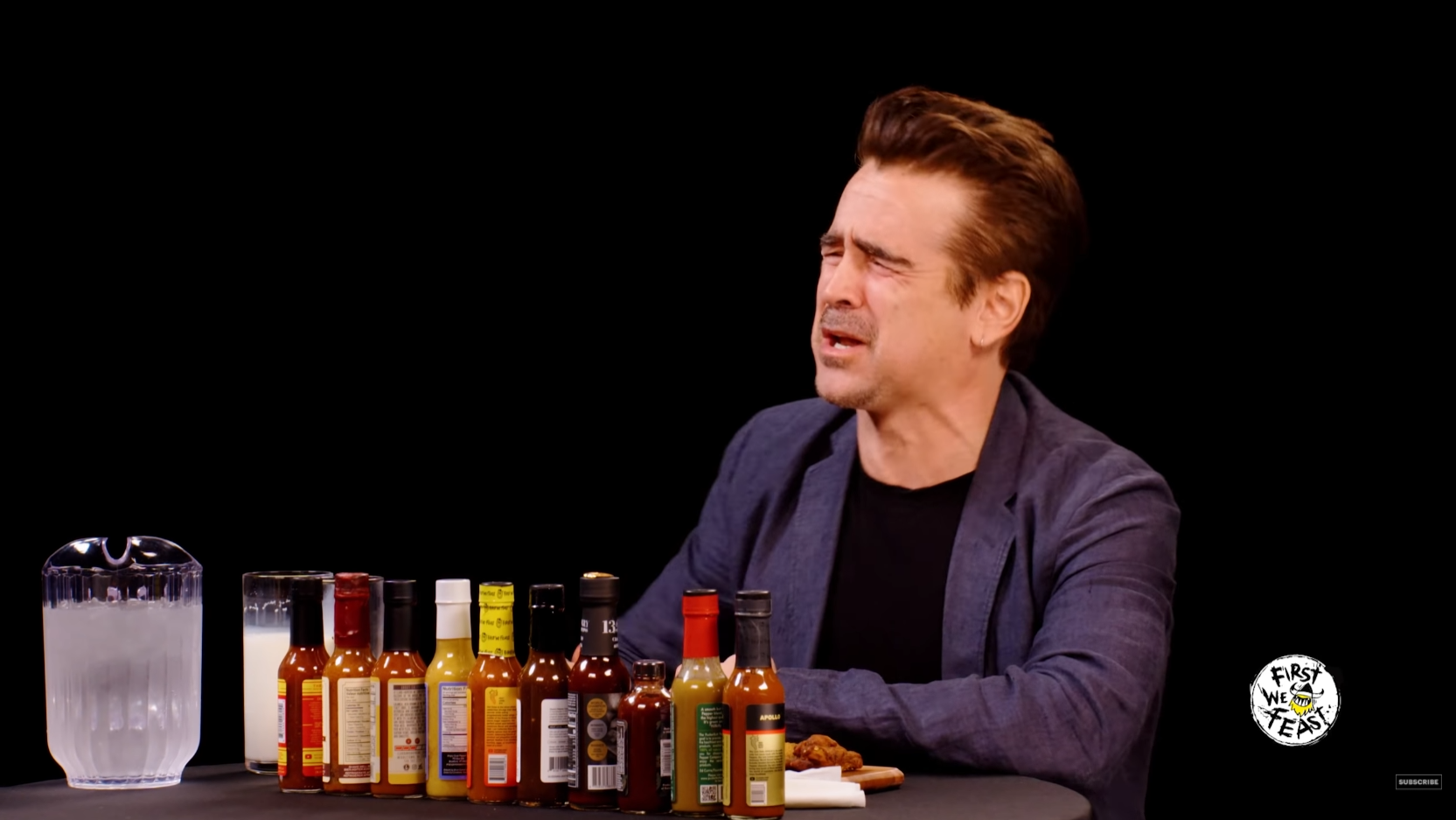 A still from the YouTube series 'Hot Ones'. Actor Colin Farrell sits in front of a row of hot sauce bottles with a pained expression on his face.