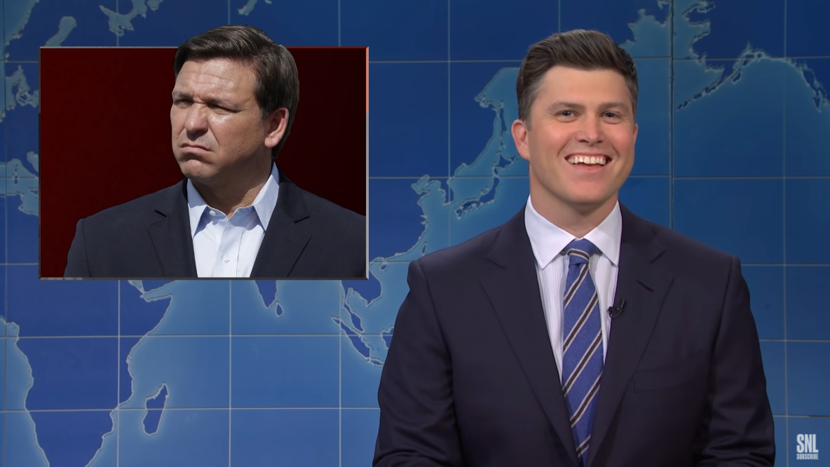 A still from Saturday Night Live. Co-host Colin Jost sits on a new set with an interposed graphic above him featuring a photo of Florida governor Ron DeSantis.