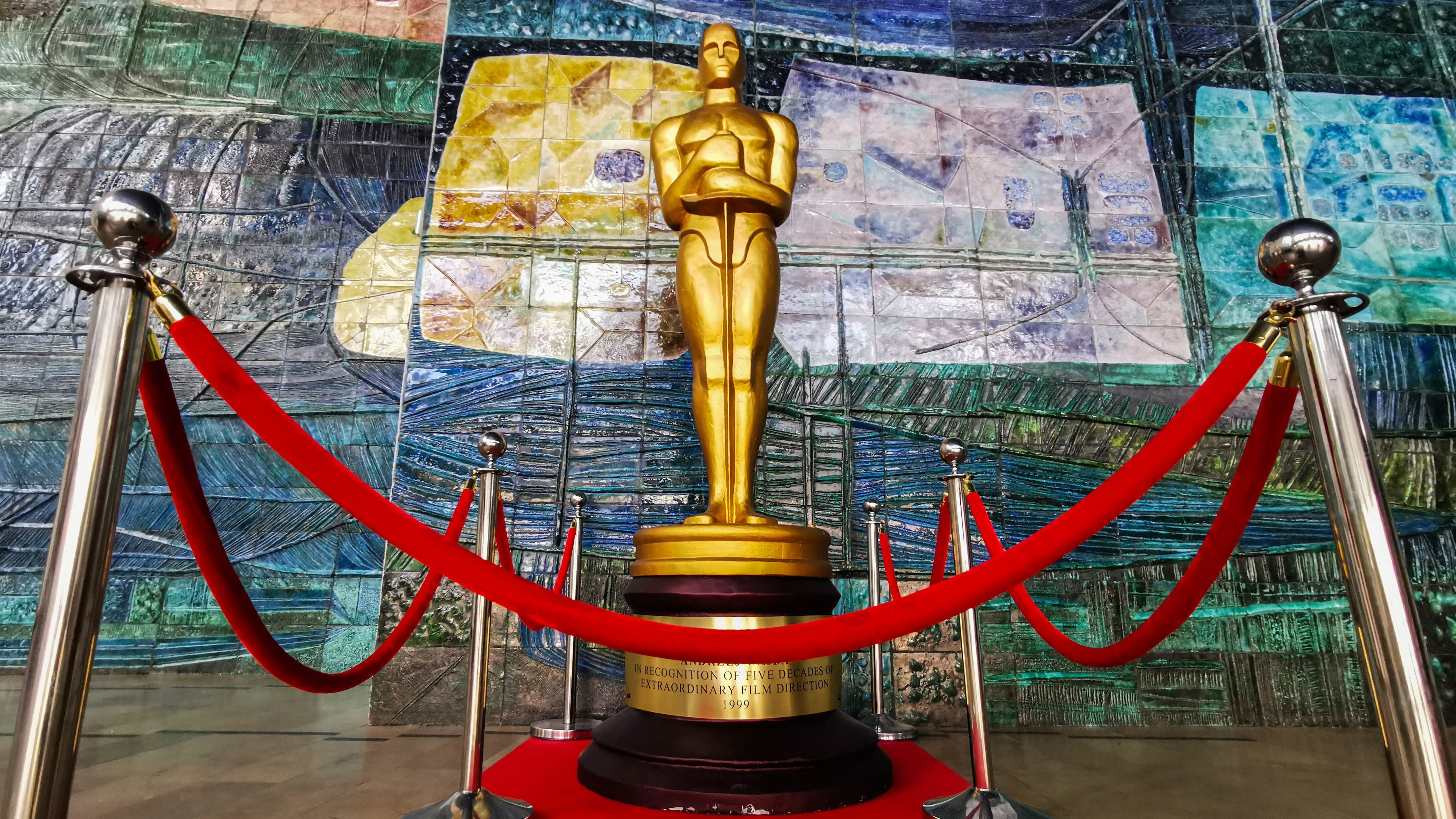 An oversized replica of the Oscars statue sealed off behind velvet rope and set up in front of a colorful wall.