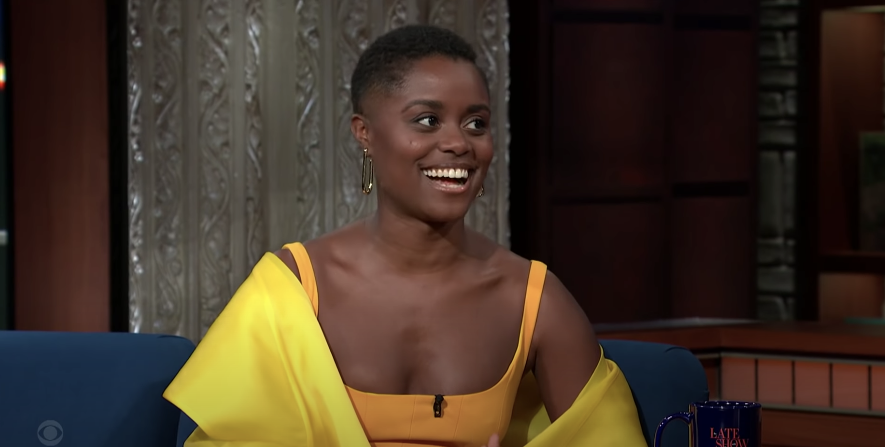 Actor Denée Benton from HBO's The Gilded Age wears a yellow dress and shawl and sits beside Stephen Colbert.