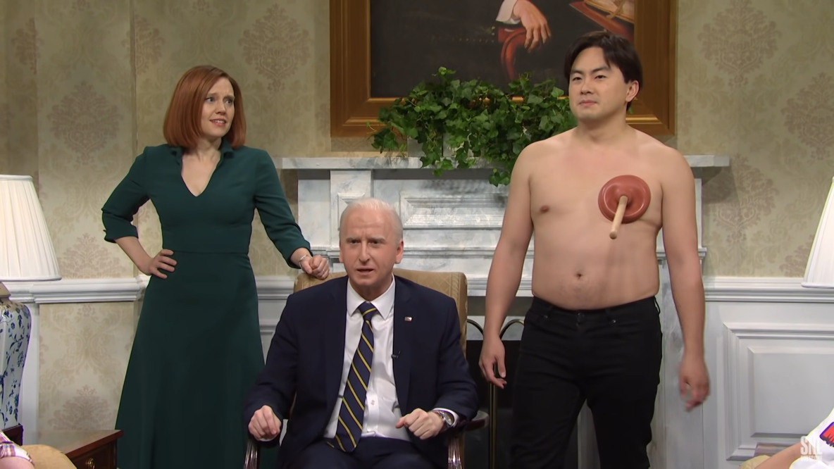 Kate McKinnon and James Austin Johnson in costume as Jen Psaki and Joe Biden, respectively, together in the White House alongside a shirtless Bowen Yang with a plunger affixed to one of his nipples.