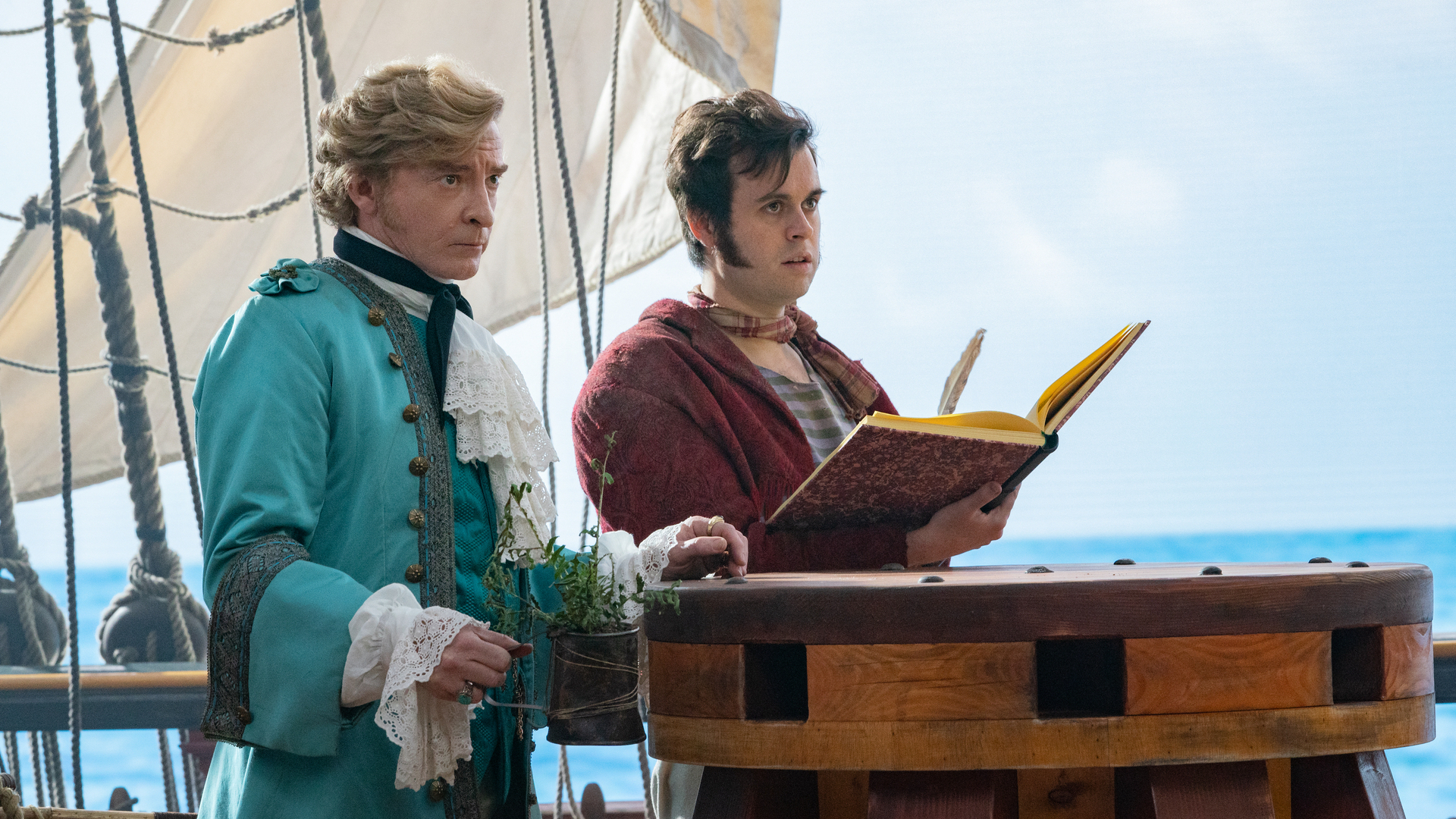 Two men: one in a fancy blue overcoat, the other in a red jacket holding a book. They stand aboard a pirate ship with the ocean behind them.