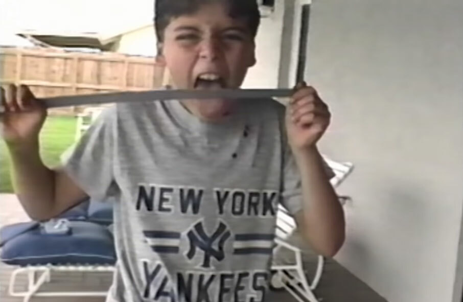 A young boy wearing a New York Yankees shirt is holding what appears to be a sword in both hands and licking its blade.