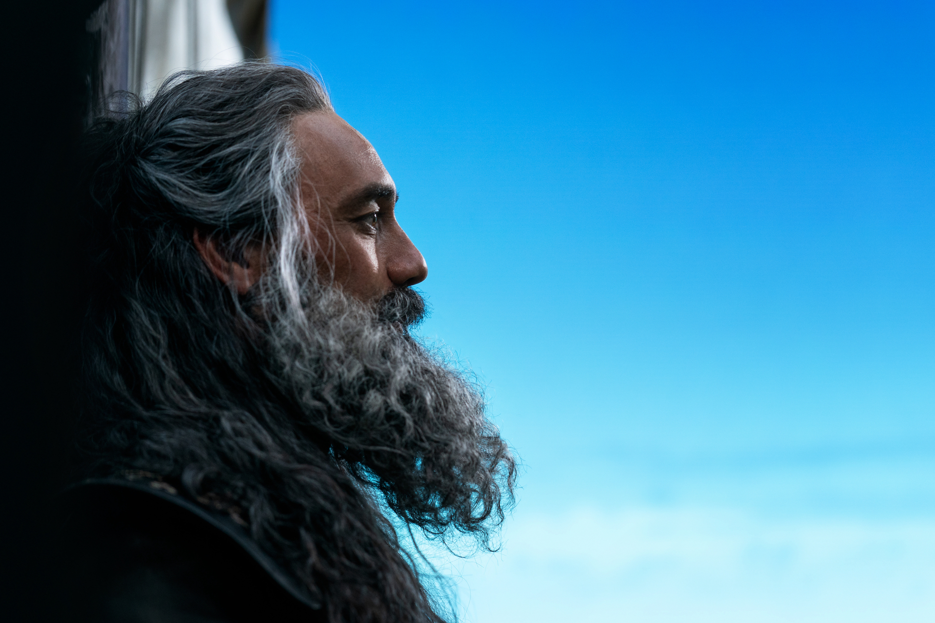 A close up on Taika Waititi as Blackbeard. He stands in profile against a very blue sky.