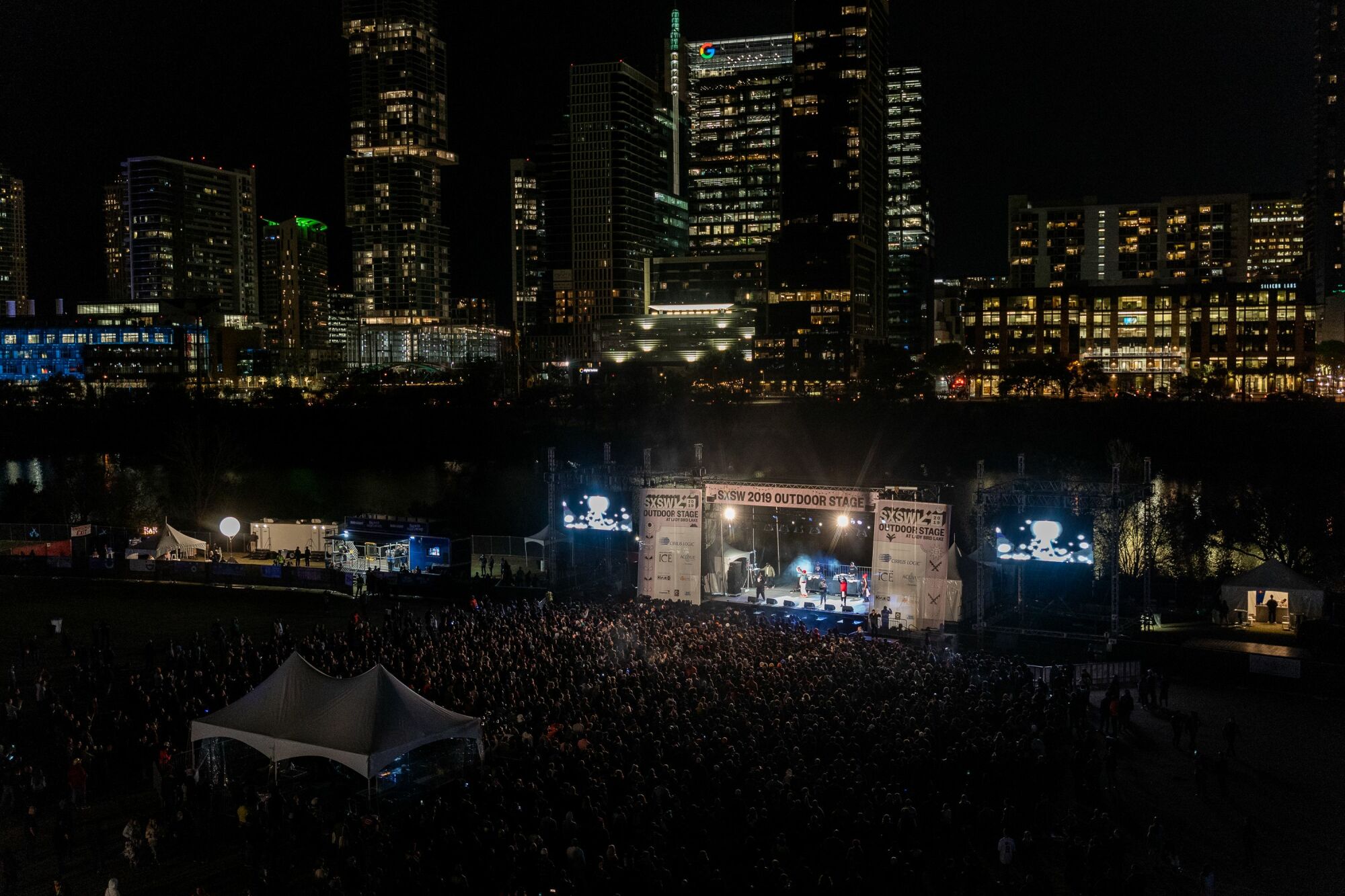 An outdoor stage with downtown buildings lit up at night.