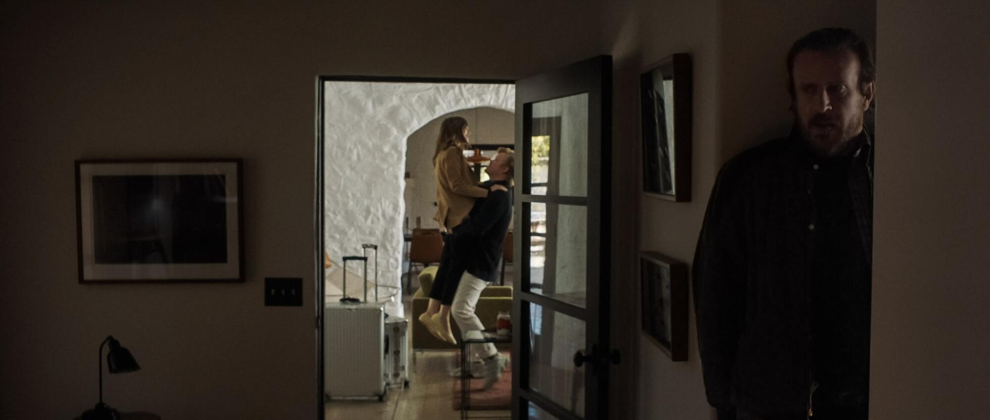 A man (Jason Segel as "Nobody") hiding behind a wall from a woman (Lily Collins as "Wife") and another man (Jesse Plemons as "CEO") in a still shot of "Windfall."