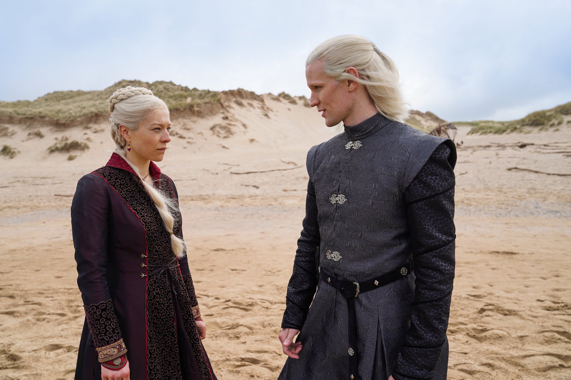 A man and a woman with platinum blonde hair face each other on a beach.