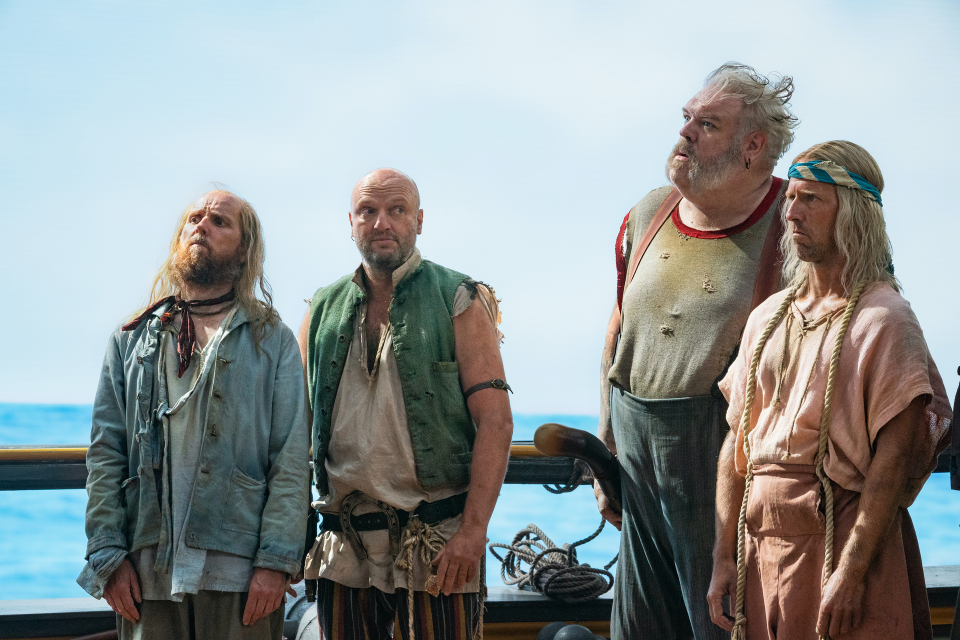 Four pirates in ragged clothes stand on the deck of their ship with the ocean behind them.