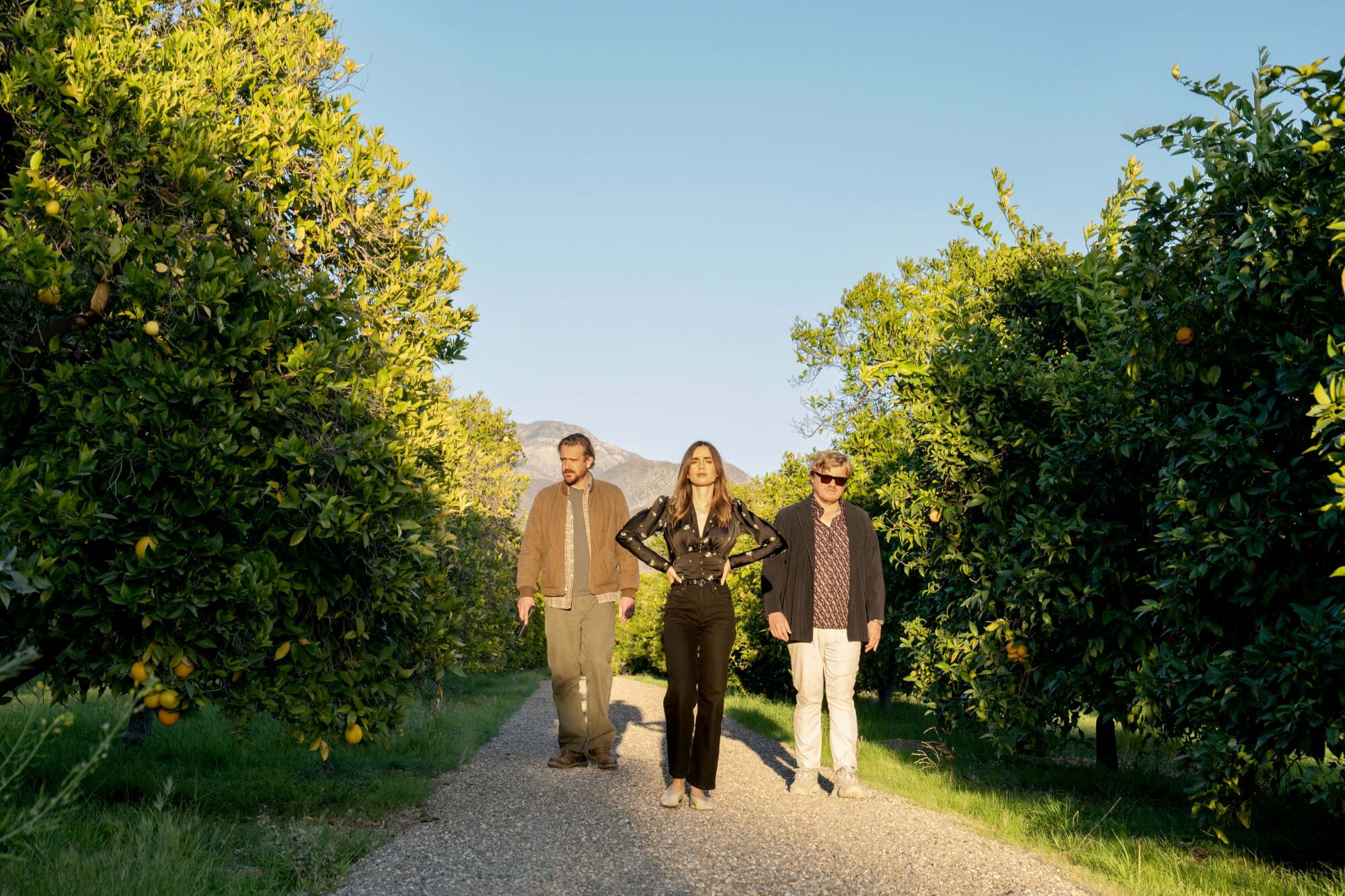 A man (Jason Segel as "Nobody"), a woman (Lily Collins as "Wife"), and another man (Jesse Plemons as "CEO") walking through an orange grove in "Windfall."