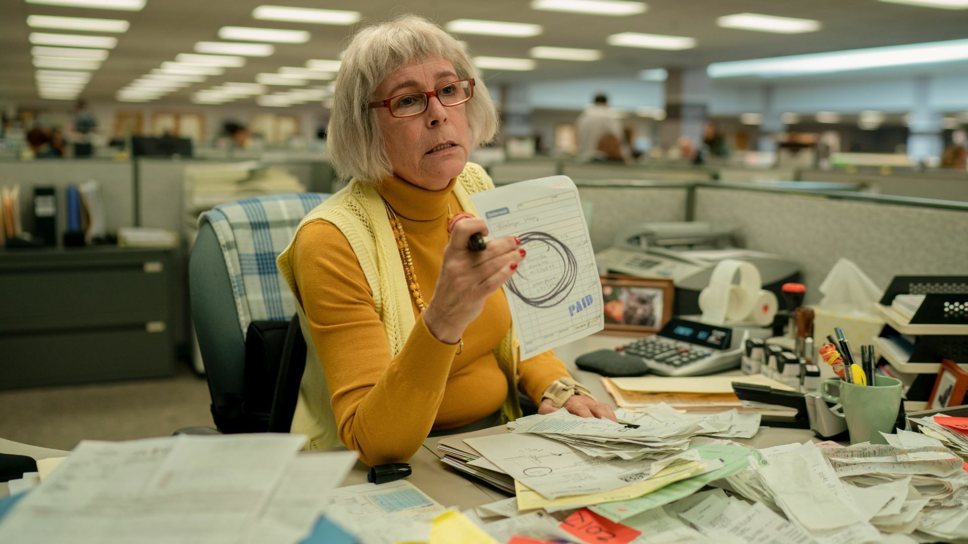 A woman in yellow sits at a office cubicle, her desk covered in receipts.