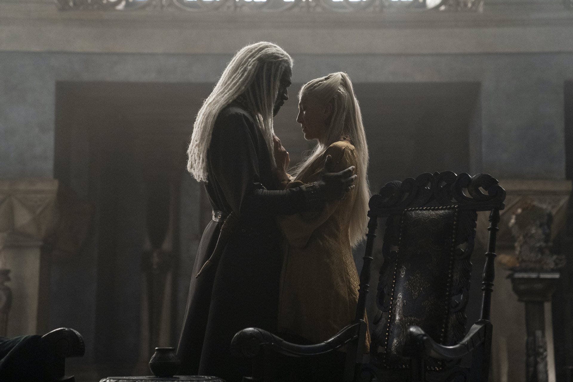 A man with white locs and a woman with long, straight blonde hair stand facing each other in a dark room, holding each other's shoulders.