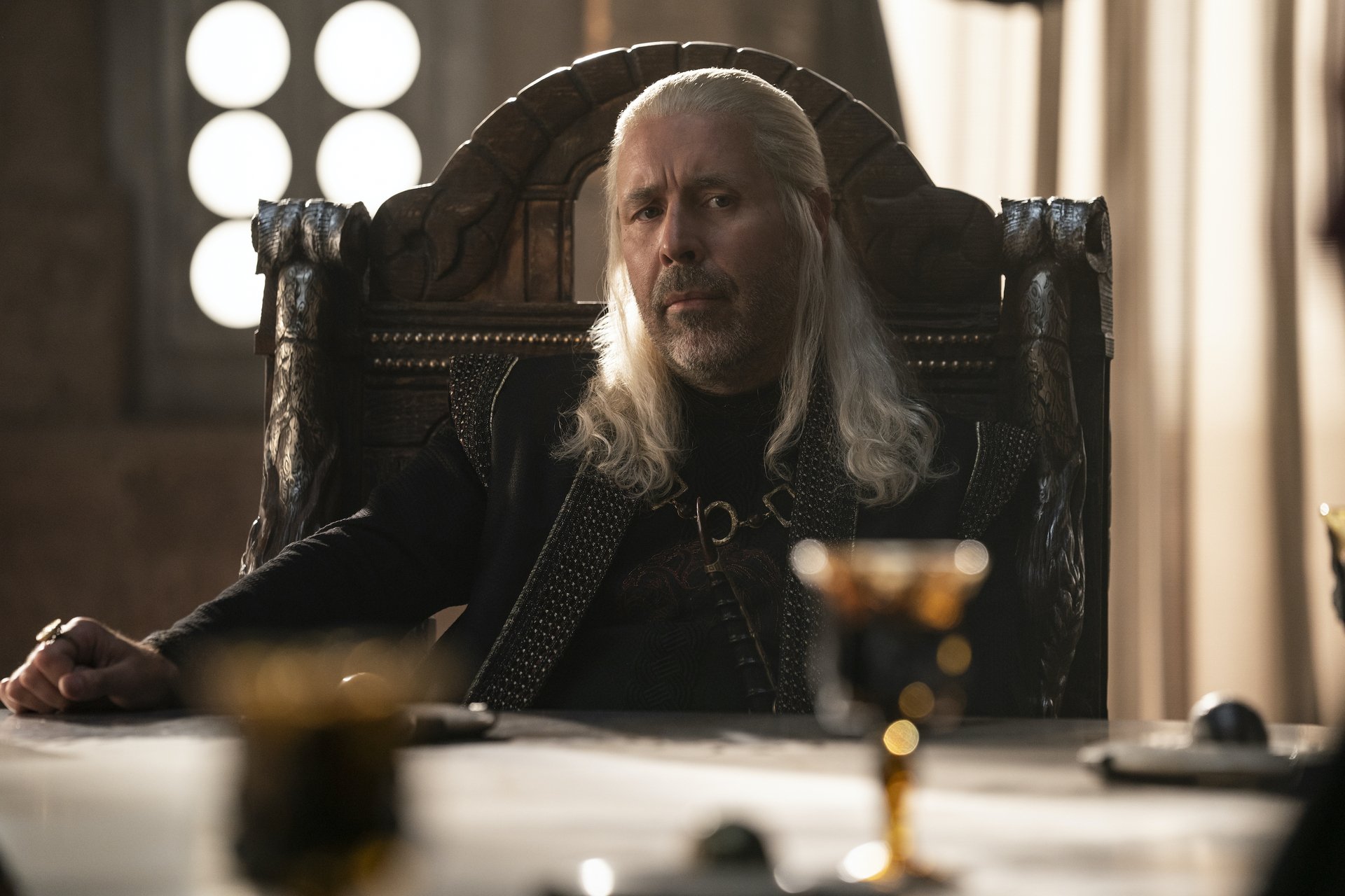 A man with long, straight white hair sits at a table in an ornately carved chair.