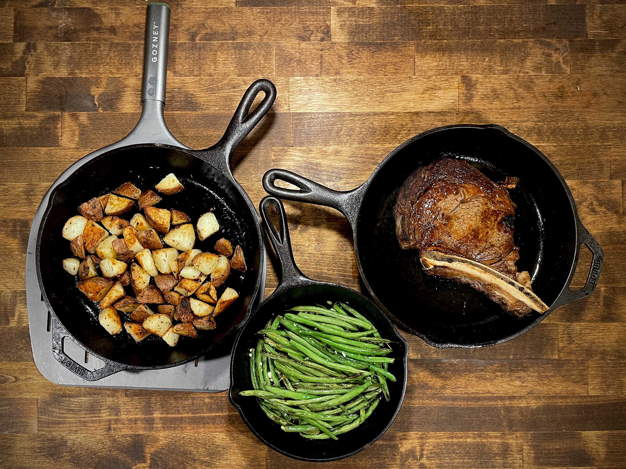 Cast iron skillets with steak, green beans, potatoes on a wood butcher block
