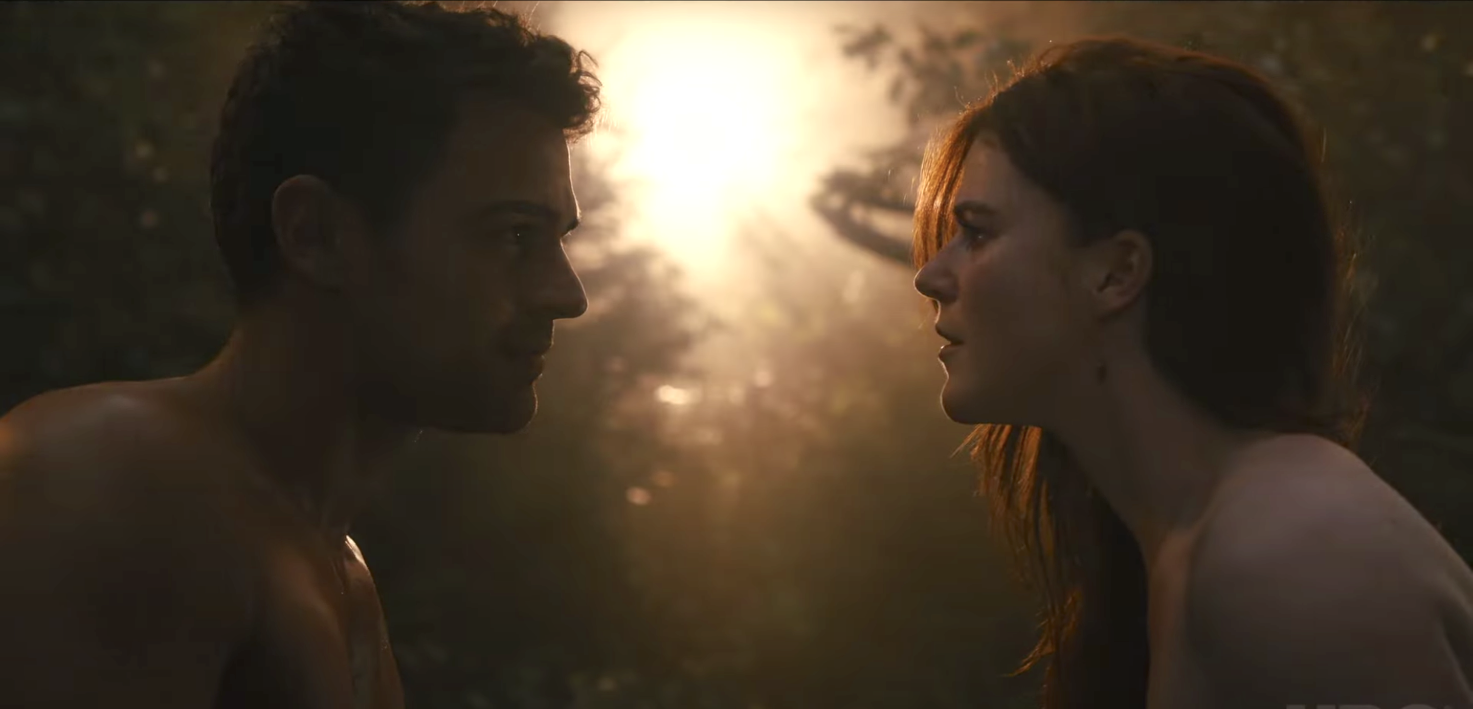 Medium-close still of a naked man and women looking at each other in a forest clearing; still from