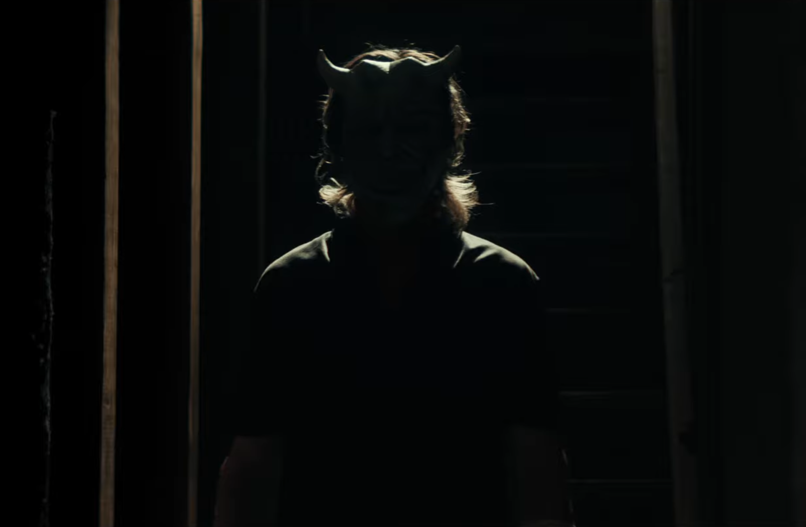 A human form stands in shadow, silhouetted by a faint light from an indistinct source. The person appears to be wearing some kind of mask over their face.