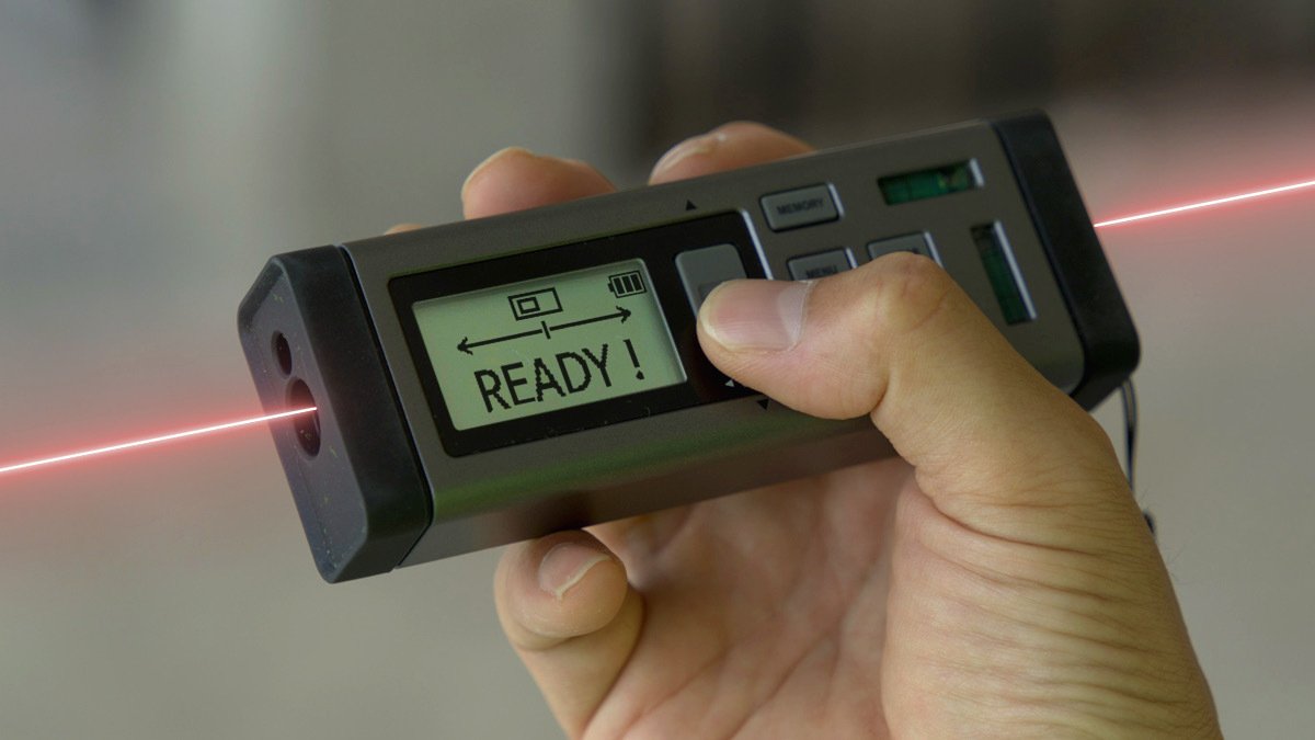 Hand holding laser distance measurer emitting red beam of light with screen that says ready