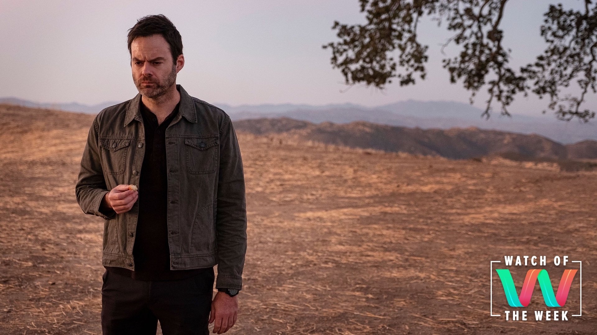 A man with dark hair and clothes stands alone in the desert, finishing the last bite of something in his hand; still from