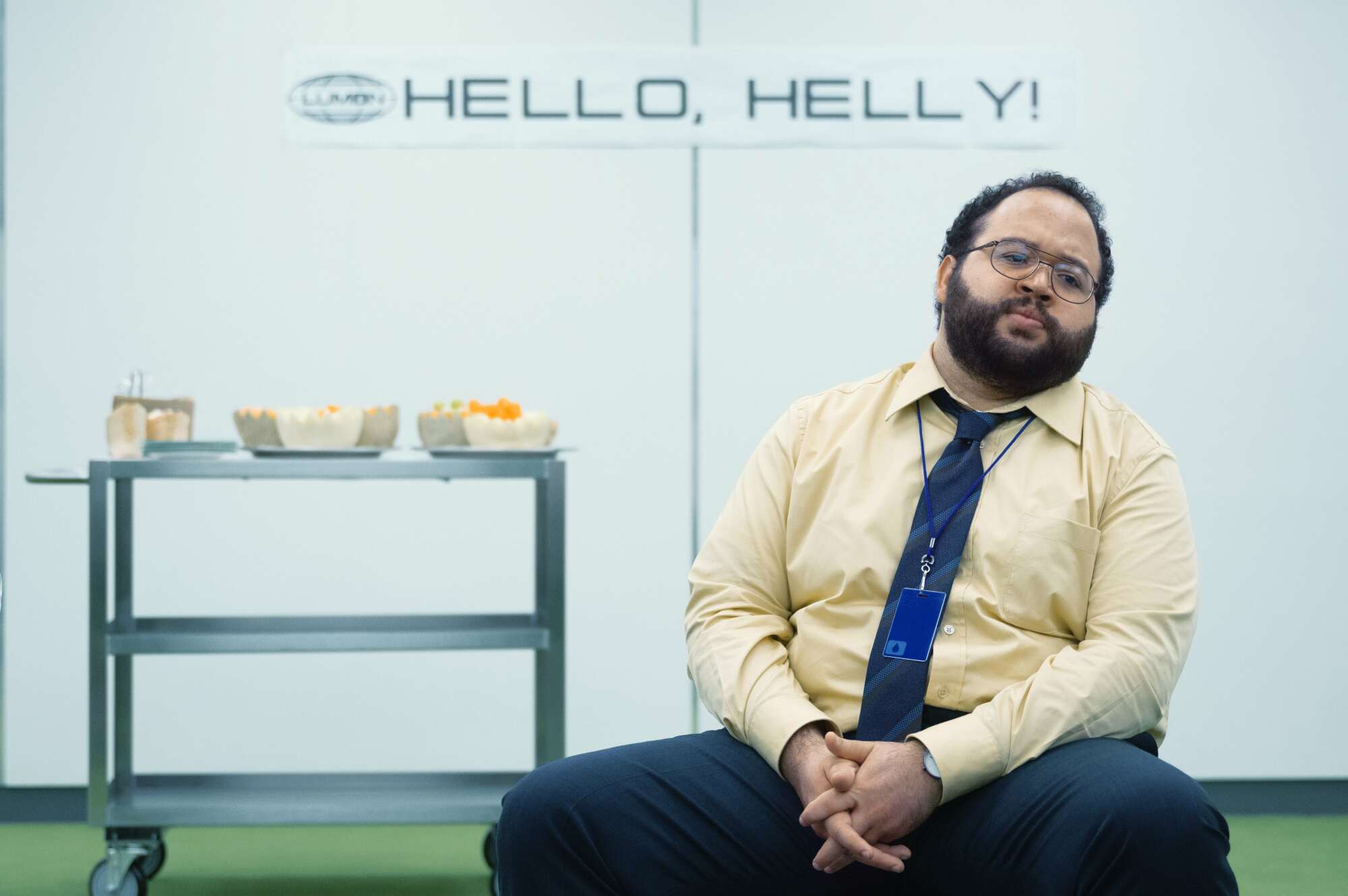 A man (Zach Cherry as Dylan in "Severance") sitting in front of a rolling cart with honeydew and cantaloupe melon.