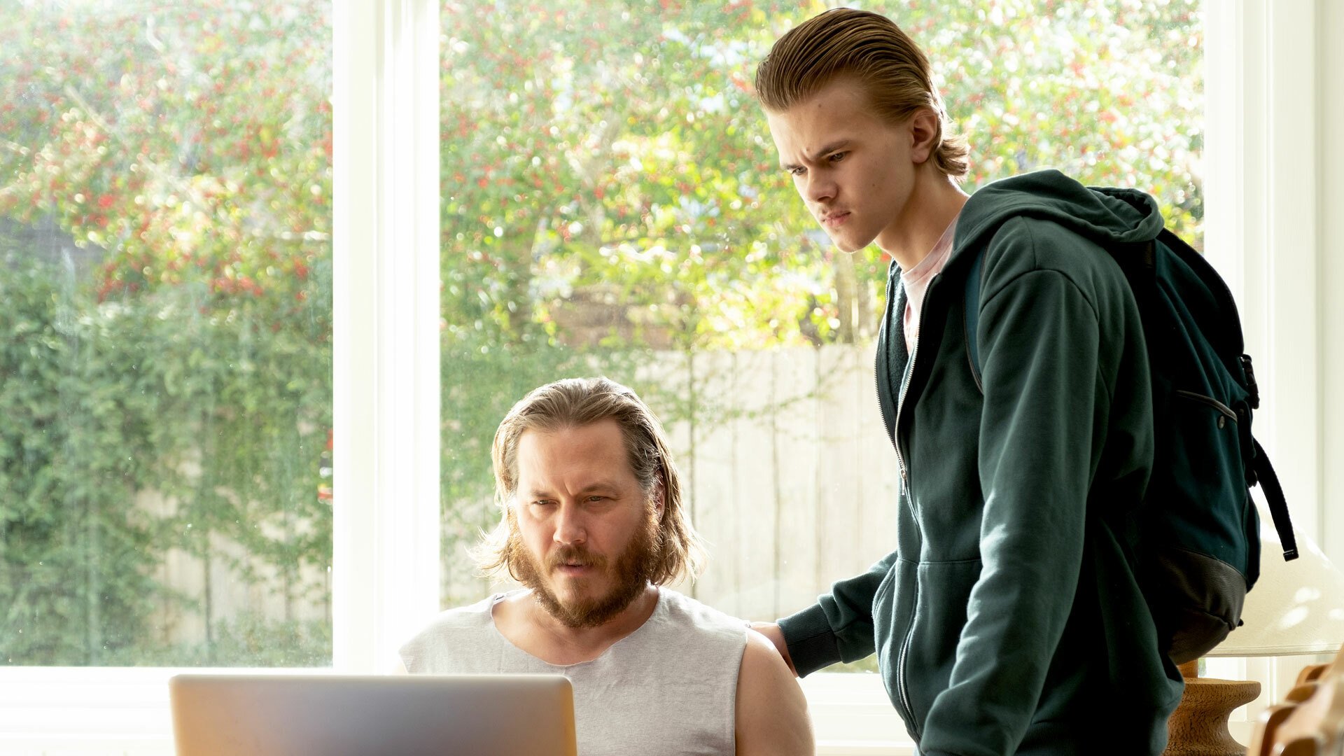 A still from Peacock's "Killing It." Brock sits at a table looking at a laptop screen while Corby stands next to him, also looking at the screen.