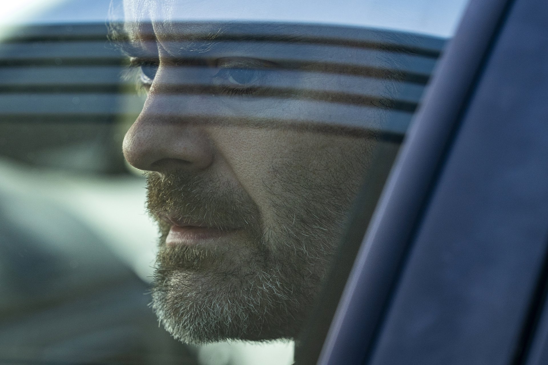 Extreme close up of a disheveled, serious man with dark and graying facial hair, staring out of a car window; still from "Barry."