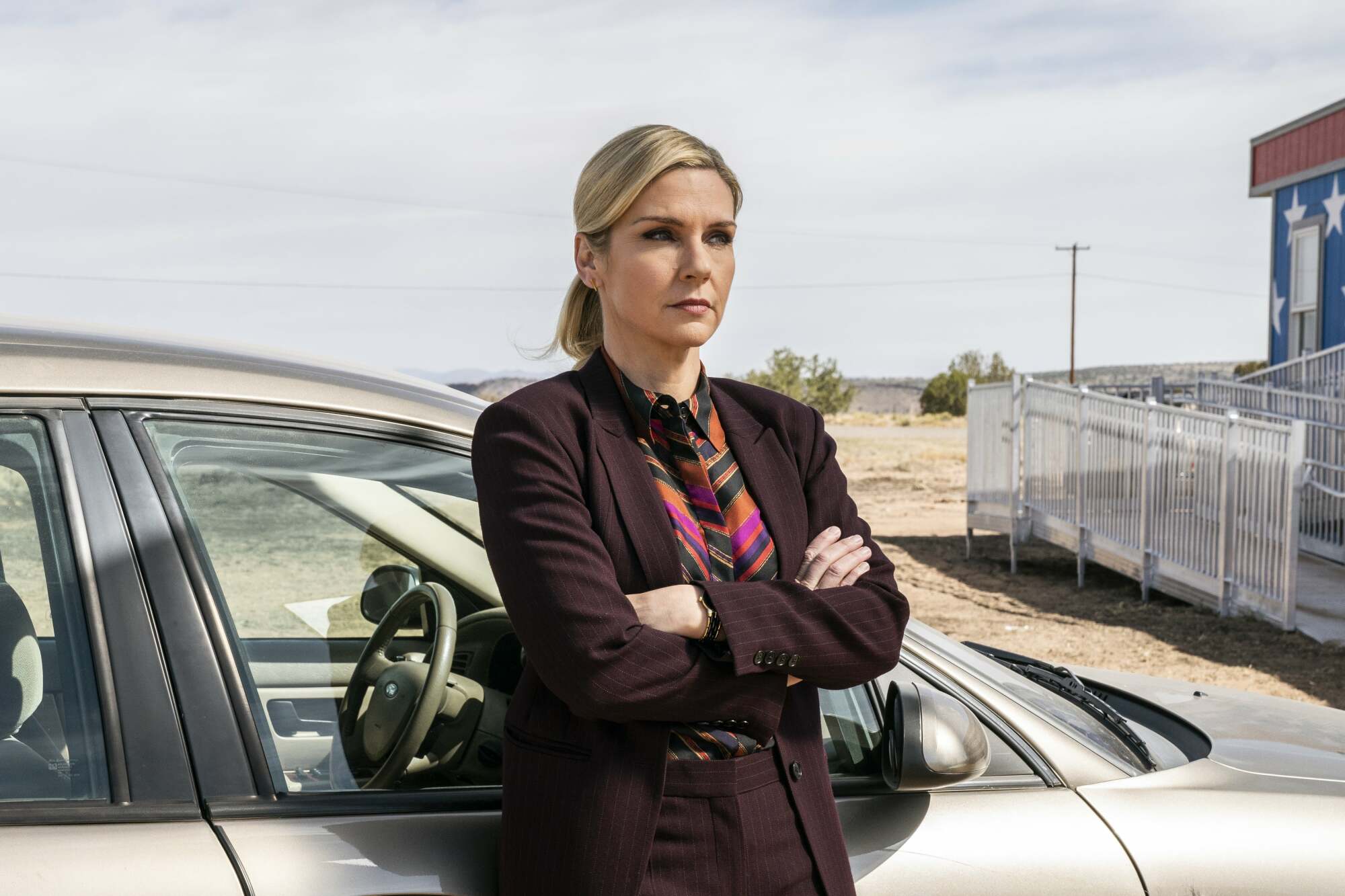 A woman (Rhea Seehorn as Kim Wexler on "Better Call Saul") leaning against a silver car with her arms crossed..