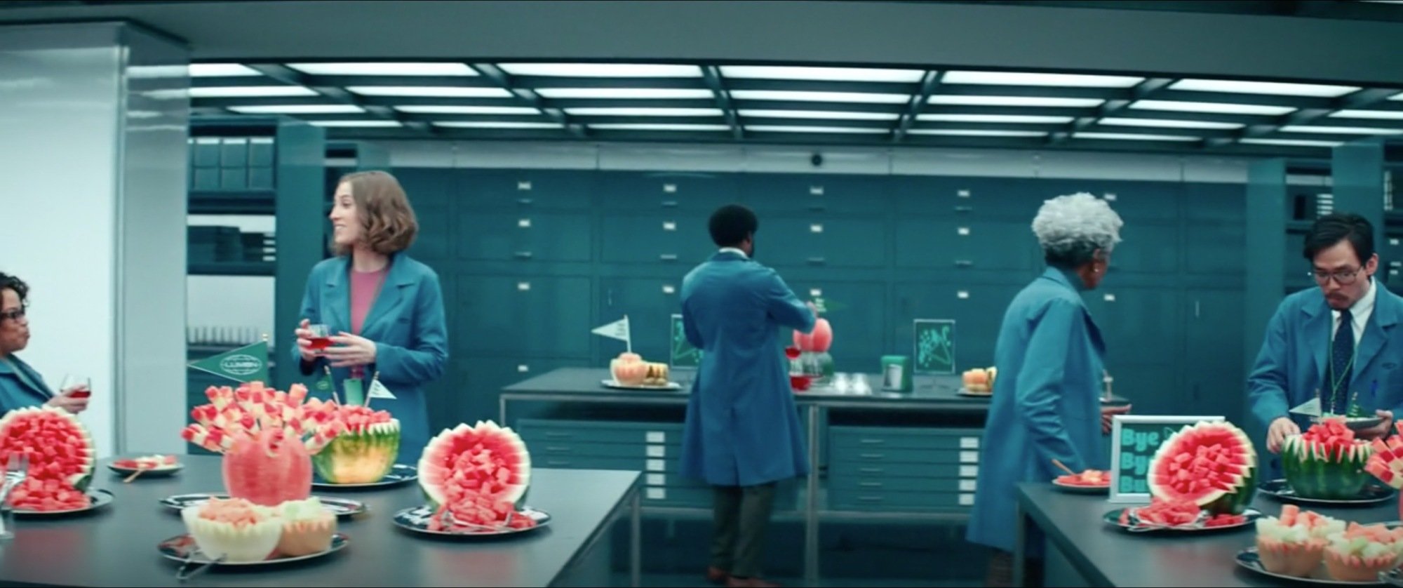 A workspace in "Severance" containing several workers and three tables with fruit bowls on them.