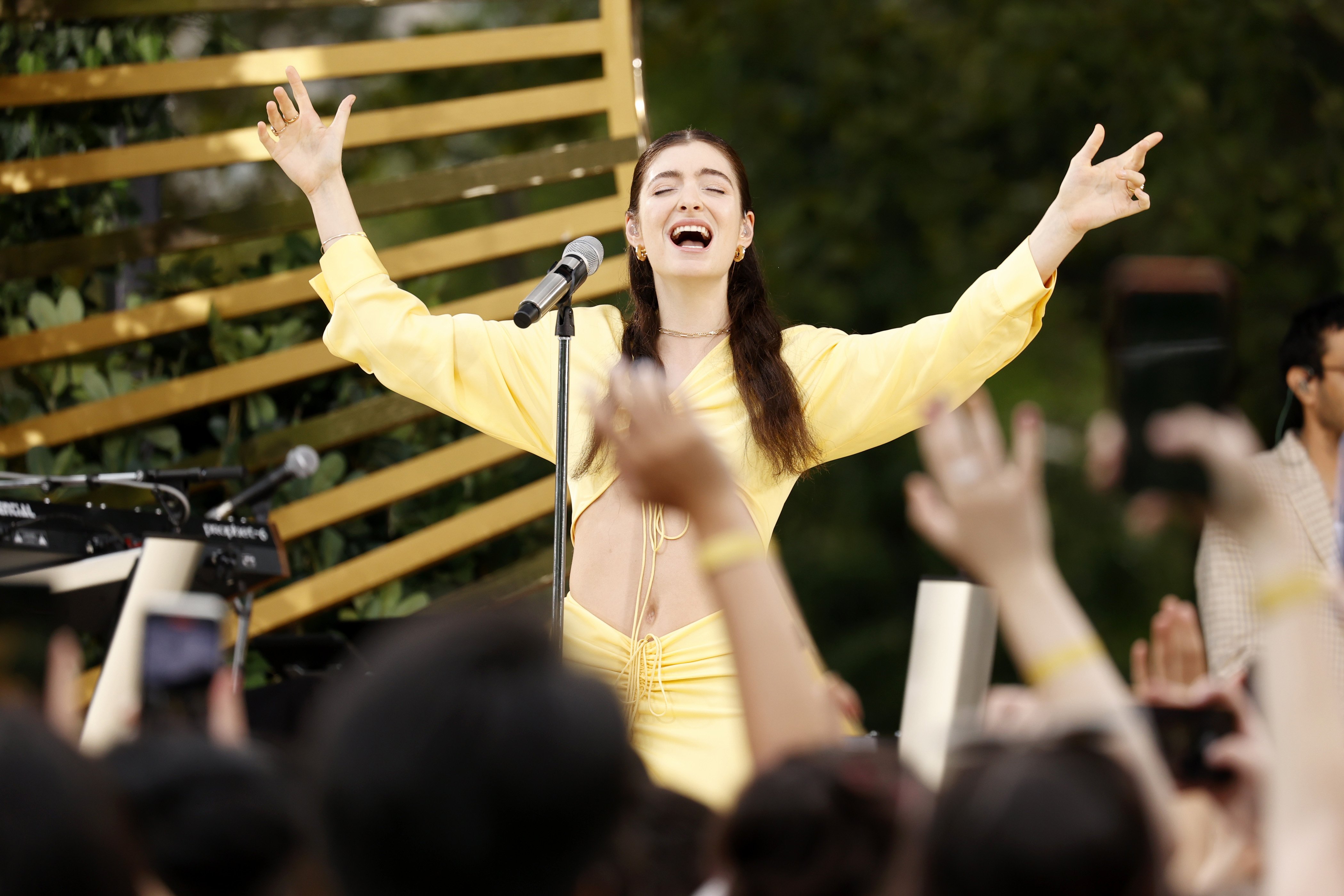 Popstar Lorde singing in a yellow dress with her arms raised above her head.