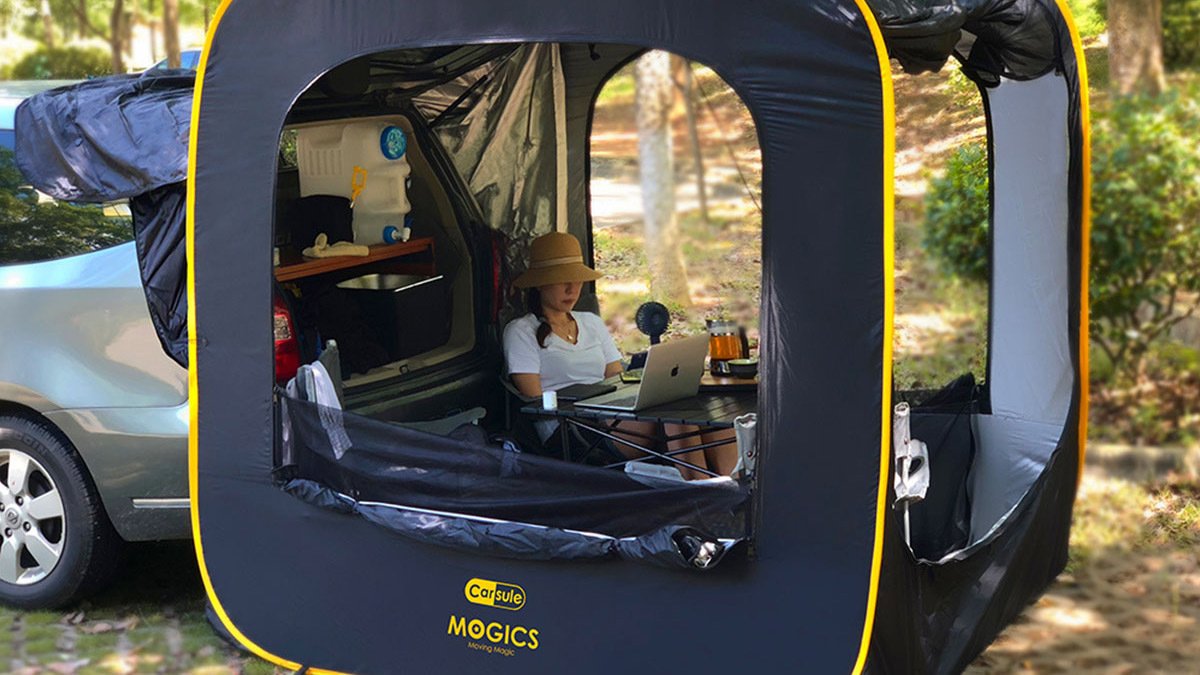 Black cubic tent attached to trunk of car with person inside on laptop