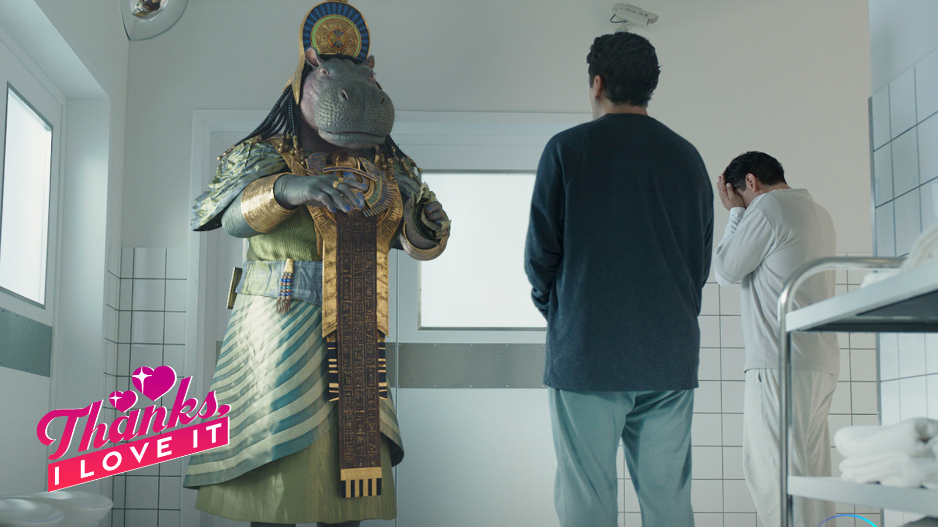 A hippo-faced goddess in Egyptian ceremonial dress speaks to two men in a hospital hallway.