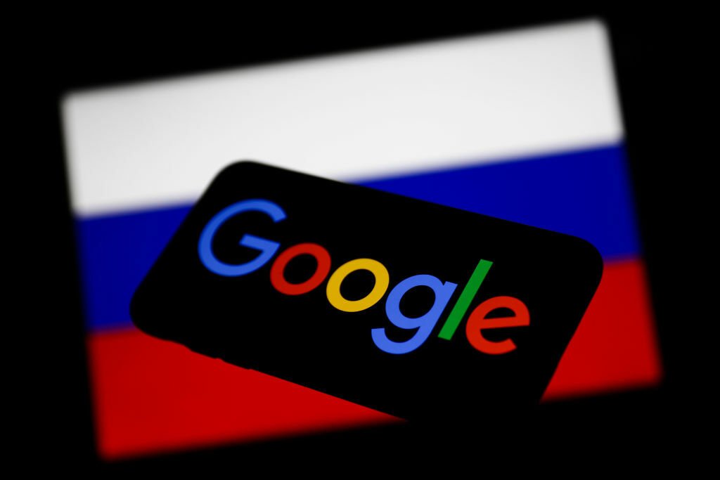 Google logo displayed on a phone screen and Russian flag displayed on a screen in the background.
