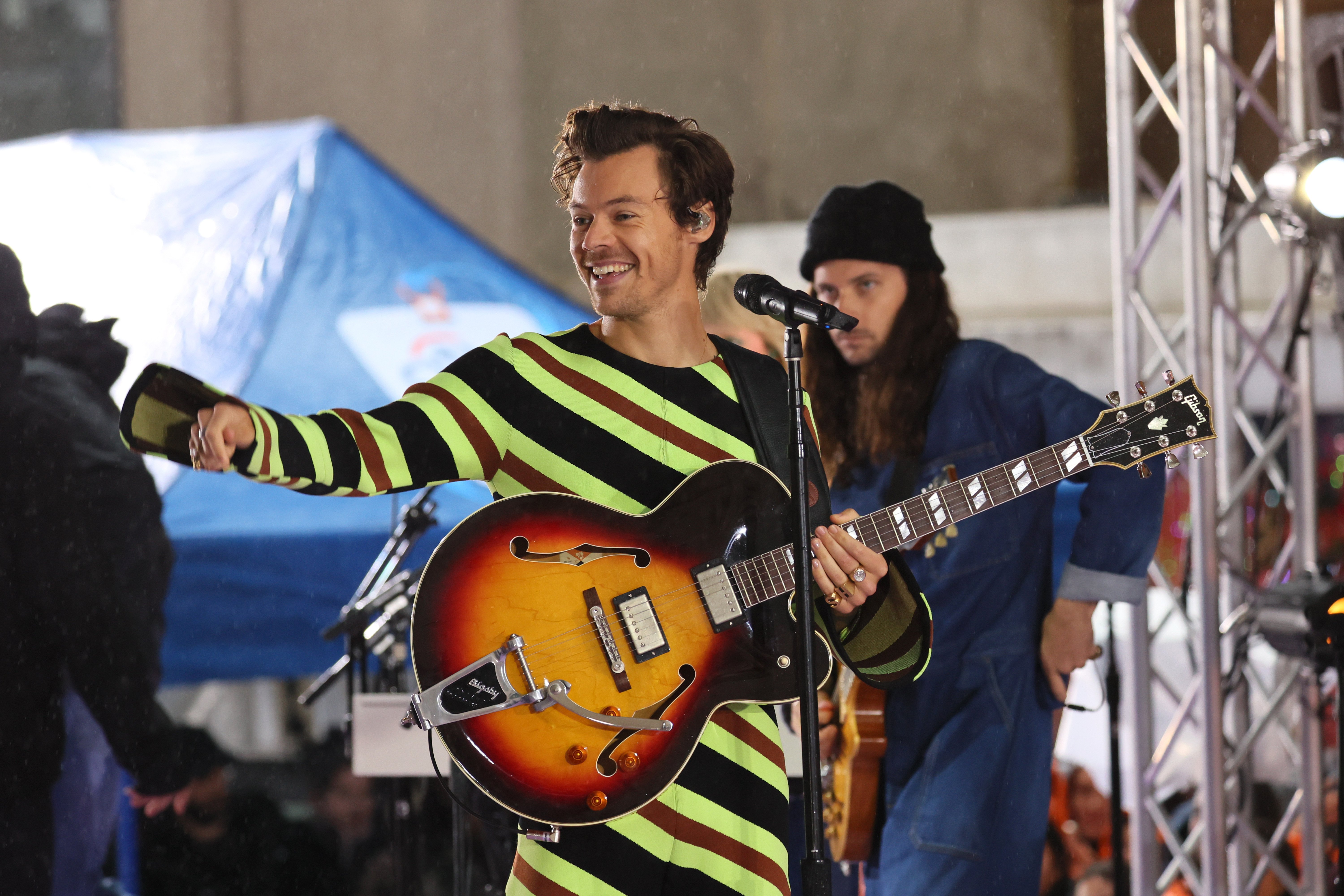 Harry Styles smiling while holding his guitar in a stripped jumpsuit on the Today Show.