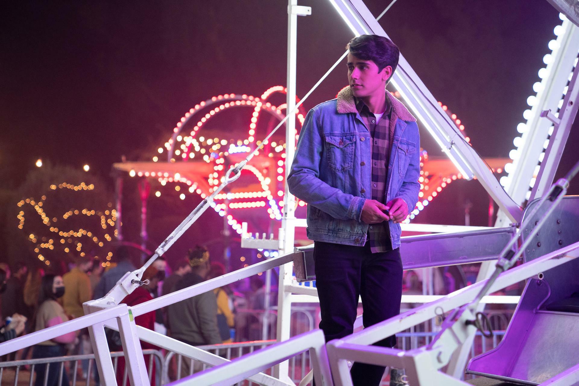 A boy in a denim jacket standing on a lit-up ferris wheel at night.