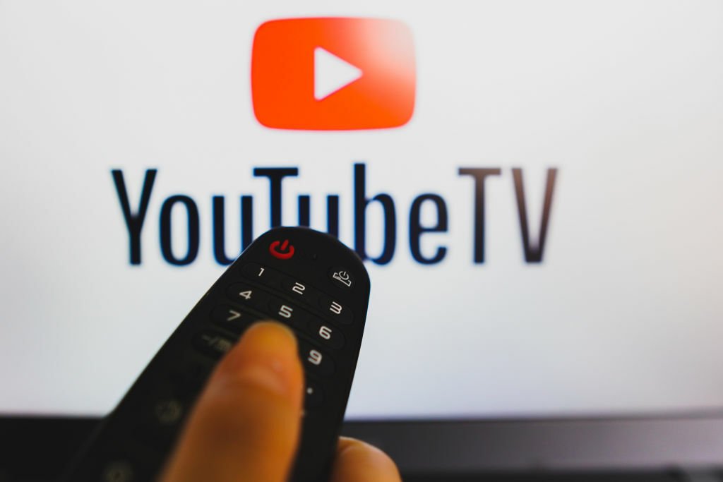 A hand holding a TV remote control in front of a YouTube logo on a TV screen.