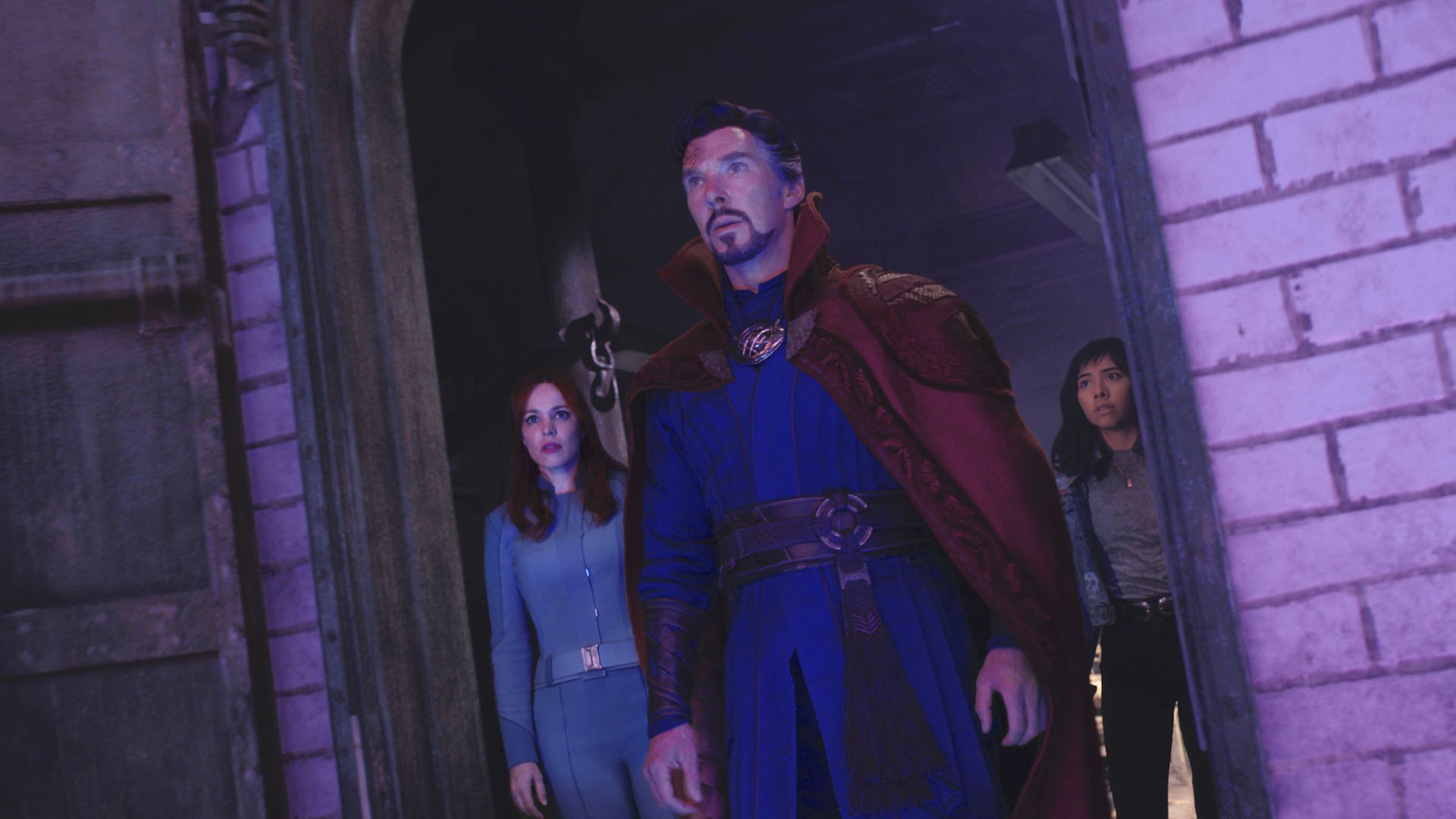 A man in a blue suit and red cape flanked by two women behind him in a doorway, all of them looking ahead at something unseen casting a purple light; still from 