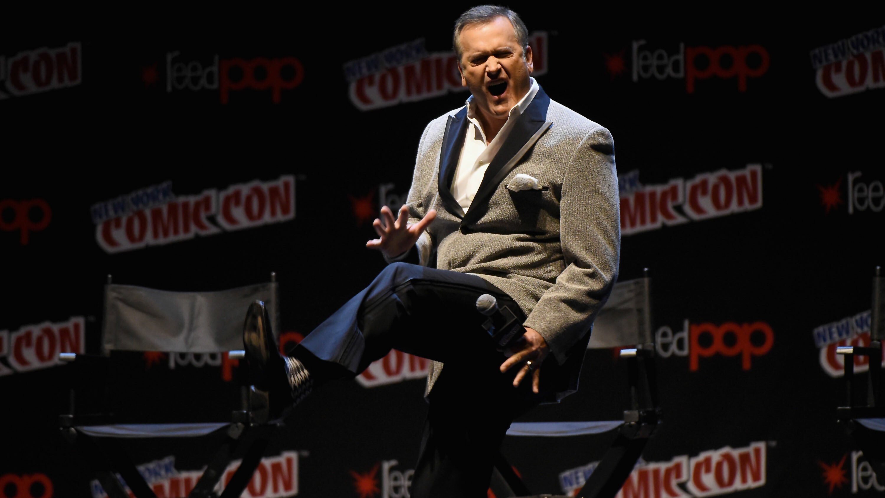 Actor Bruce Campbell kicks out one leg as he appears to shout from a stage at New York Comic-Con.