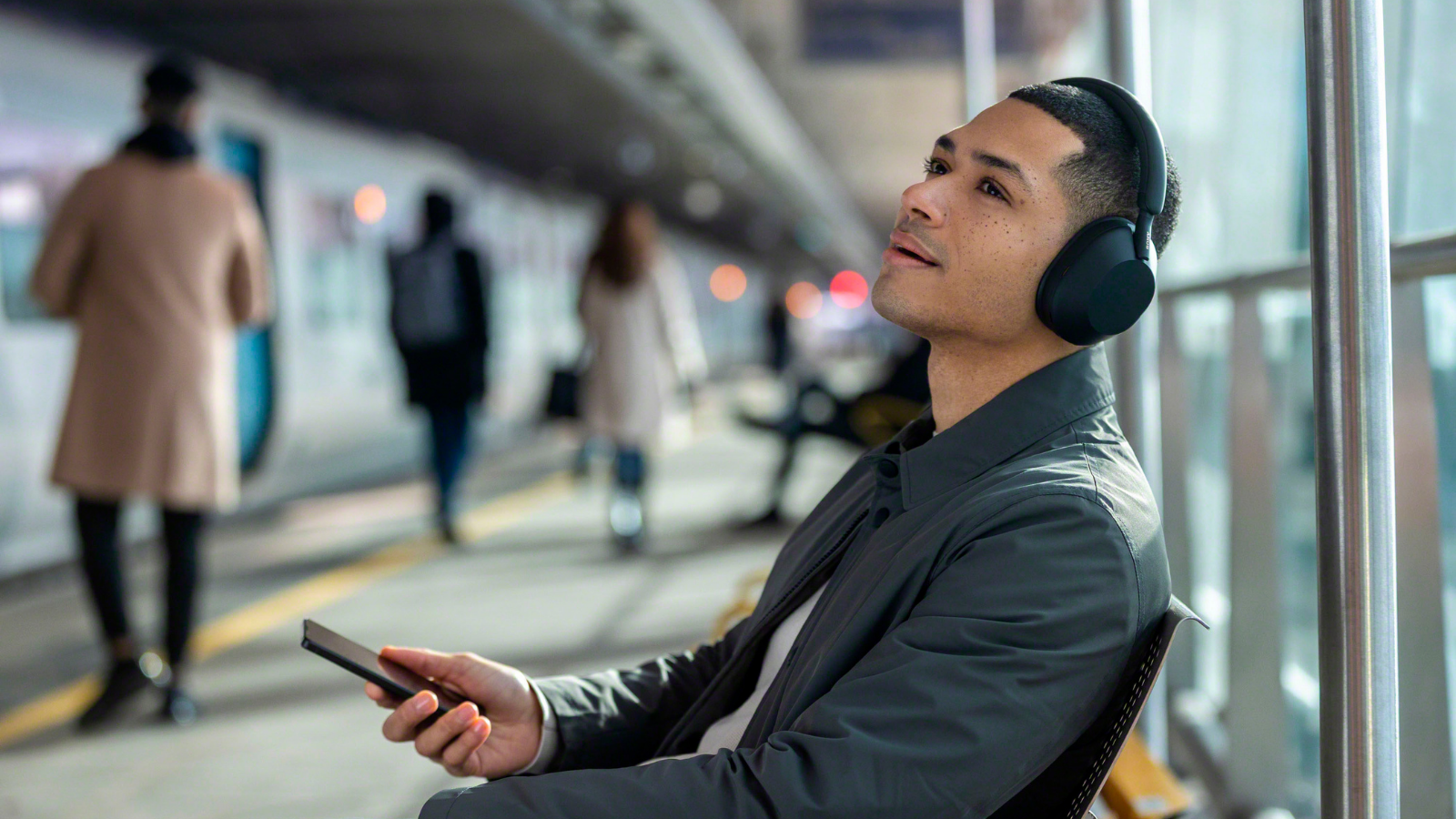 a man sitting in a subway station wearing the black Sony WH-1000XM5 Headphones while holding a phone