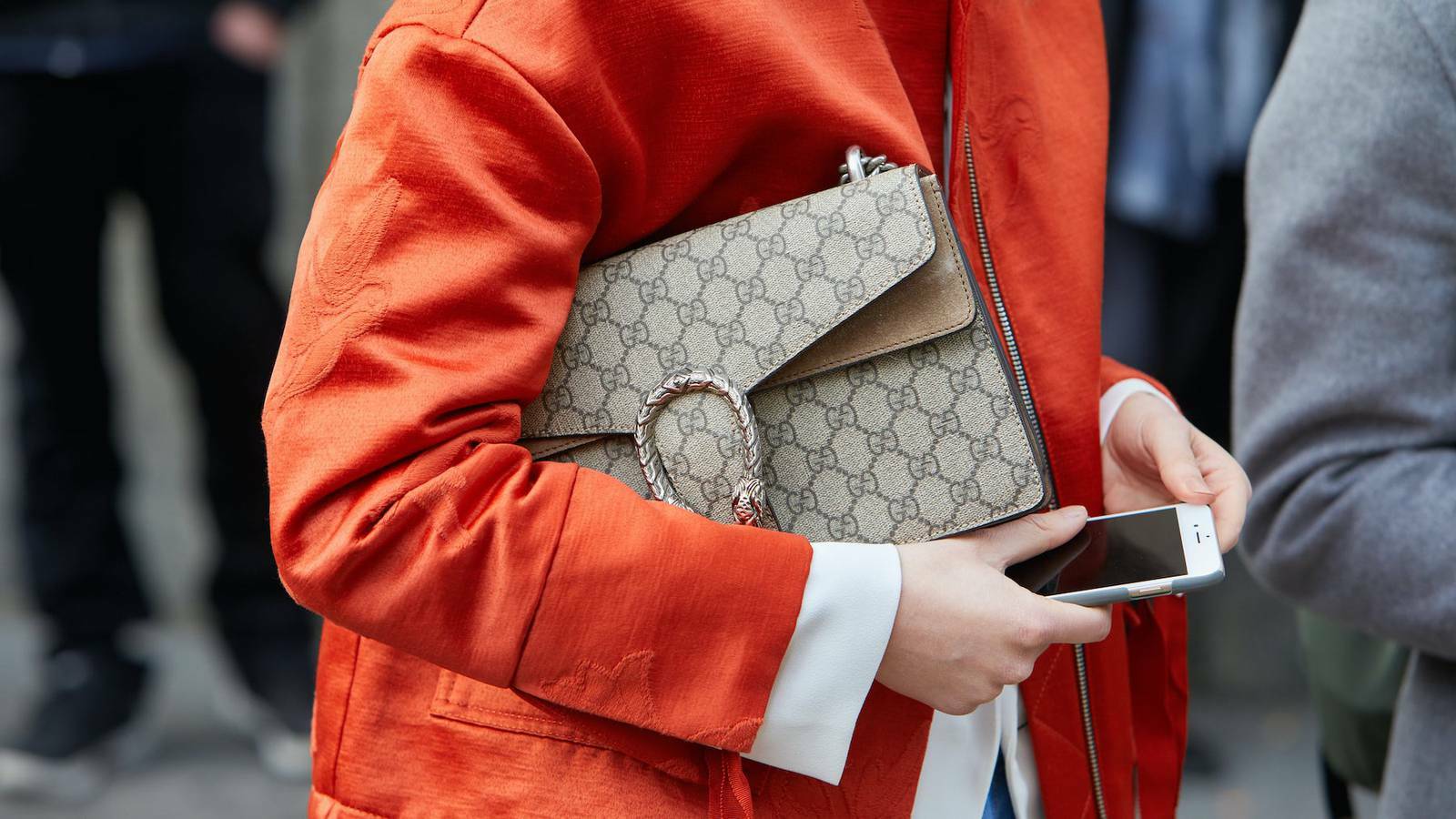 A person wearing an orange jacket, wearing a Gucci purse with the brand's logo all across it, and holding an iPhone in hand.